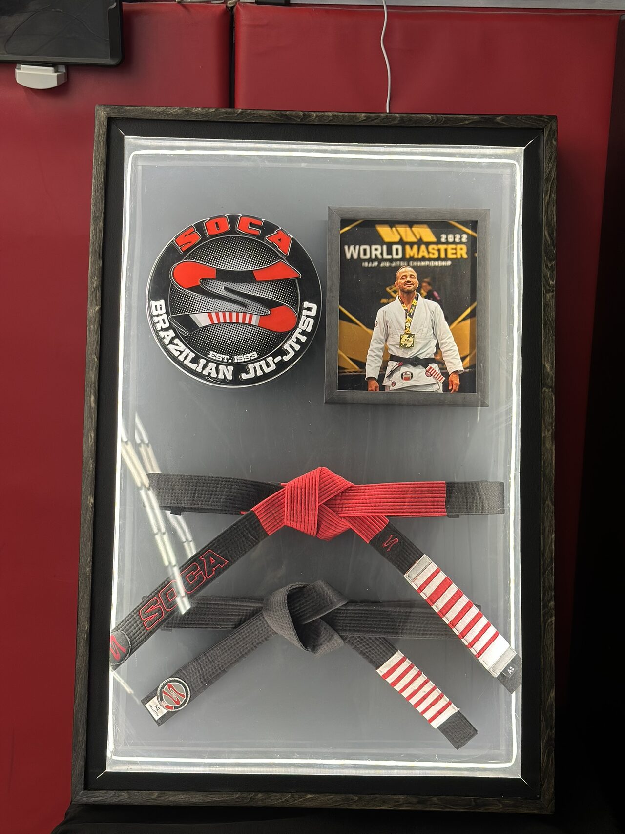 The whole display case standing upright against a red padded wall: a dark frame around a glowing panel with a round Soca logo, a framed World Master 2022 photo and two knotted belts.
