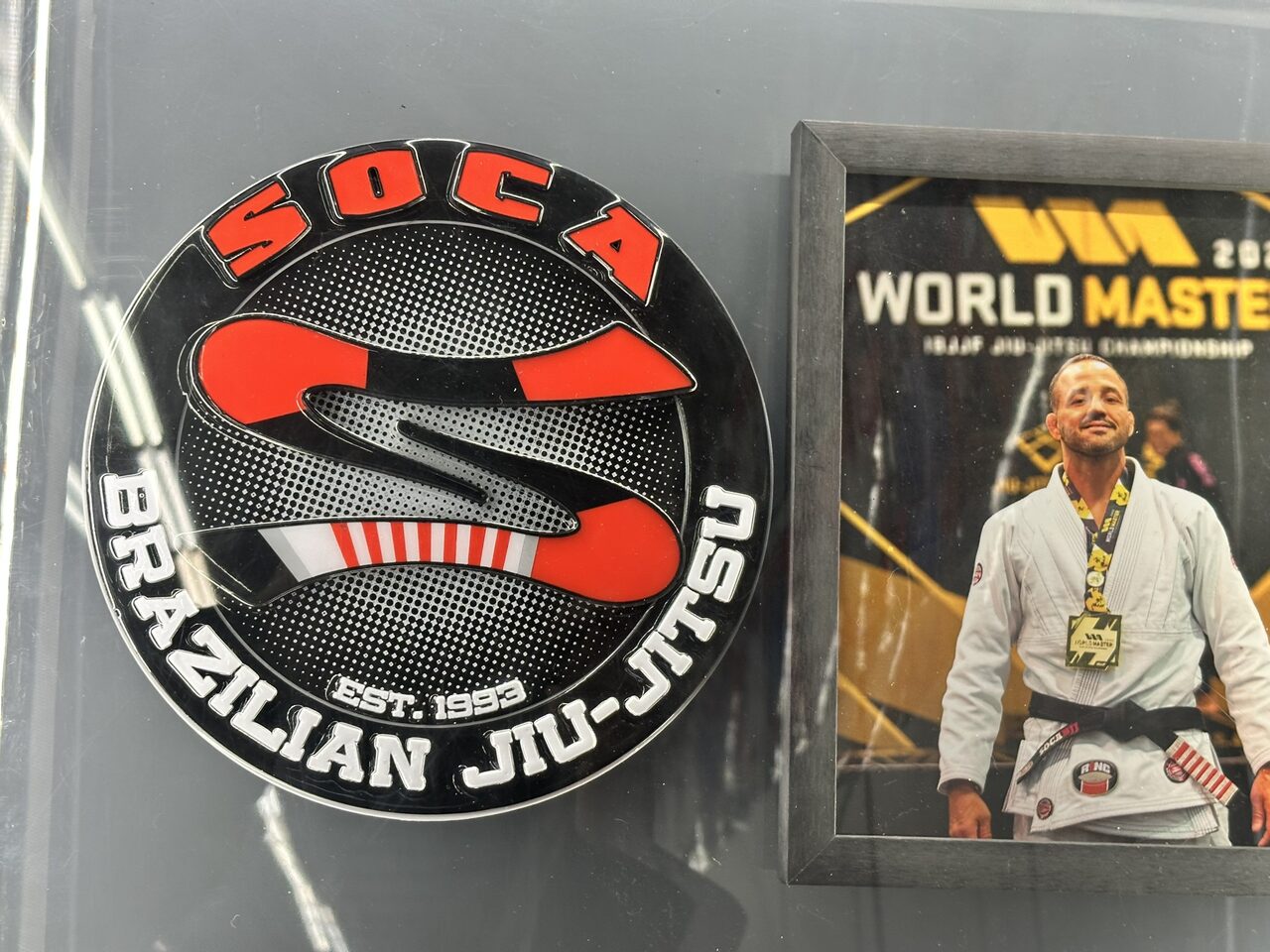 Close view of the round raised Soca Brazilian Jiu-Jitsu logo beside a framed World Master 2022 photo of a man in a white gi wearing a medal.
