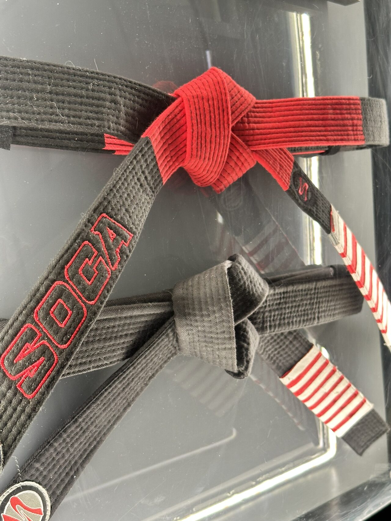 Close view through the case front of a knotted black belt with a red bar and red-and-white stripes, SOCA embroidered in red, above a second knotted belt.