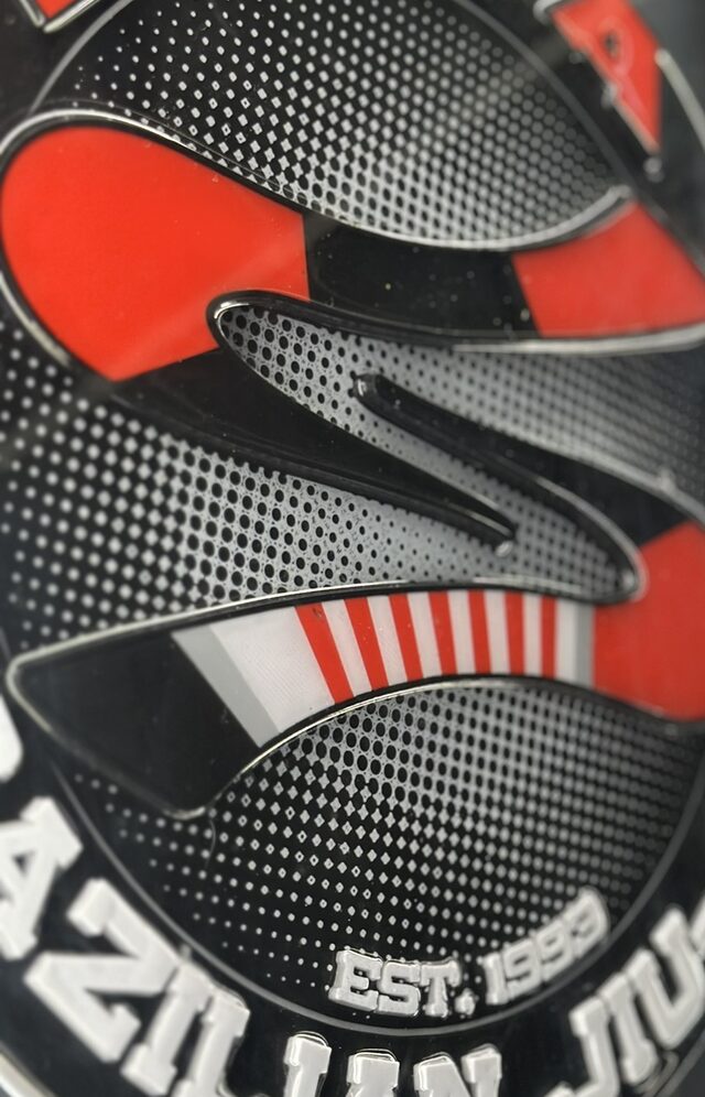 Steep side view of the round raised Soca logo in black, red and white, with halftone dots and the words EST. 1993.