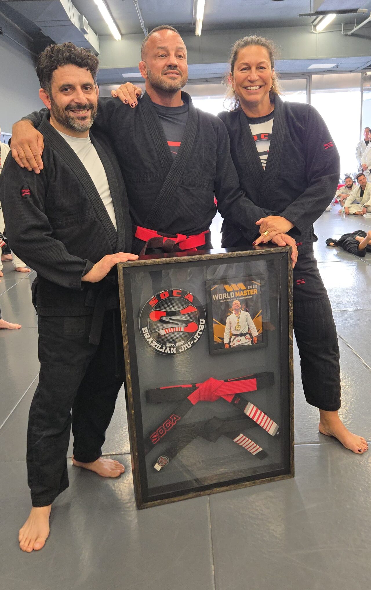 Three adults in black gis standing on the academy mats behind the finished display case, smiling, hands resting on its frame.