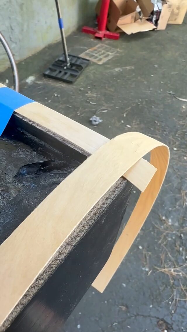 A coil of wood veneer banding held against the corner of the black frame, with blue tape holding it in place.