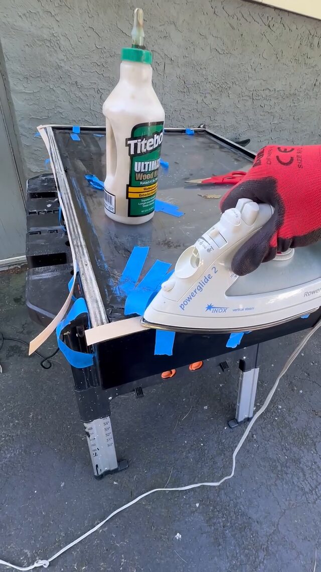 A gloved hand running a clothes iron along the taped frame edge, a glue bottle standing on the panel.