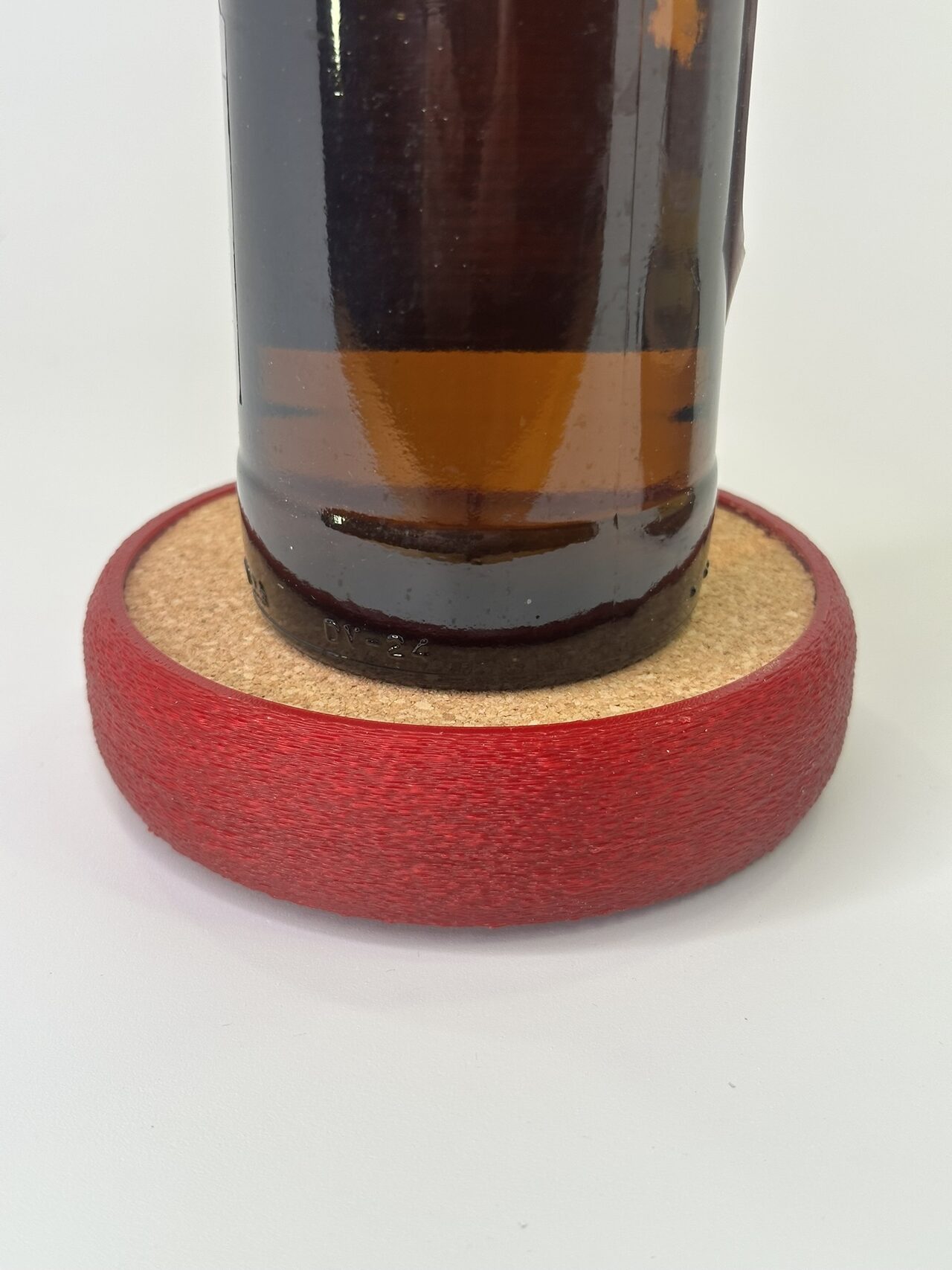 The base of a brown glass bottle sitting on the cork top of a red textured coaster, white background