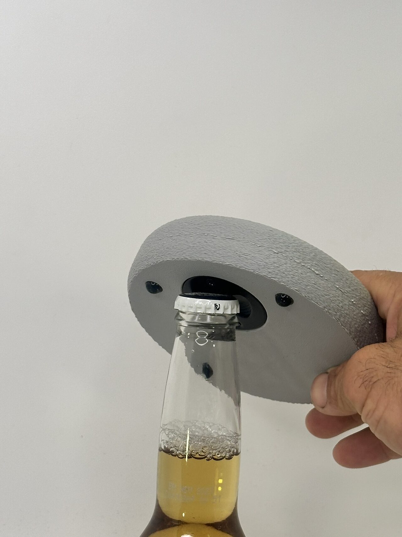 A hand levers a grey coaster against the cap of a glass bottle, using the opener in its underside