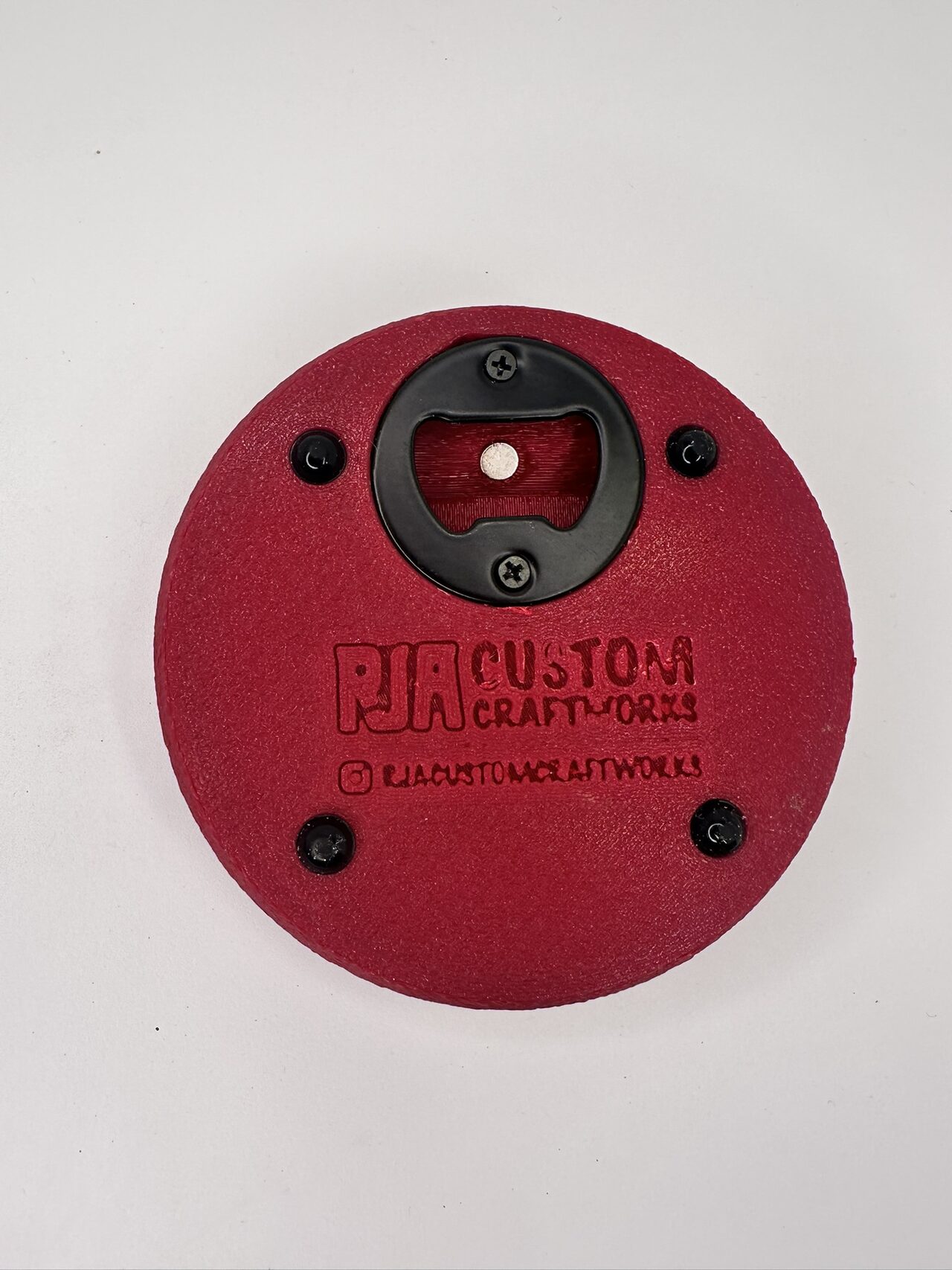 Underside of the red coaster from above: black metal bottle opener held by screws, with RJA Custom Craftworks raised in the plastic
