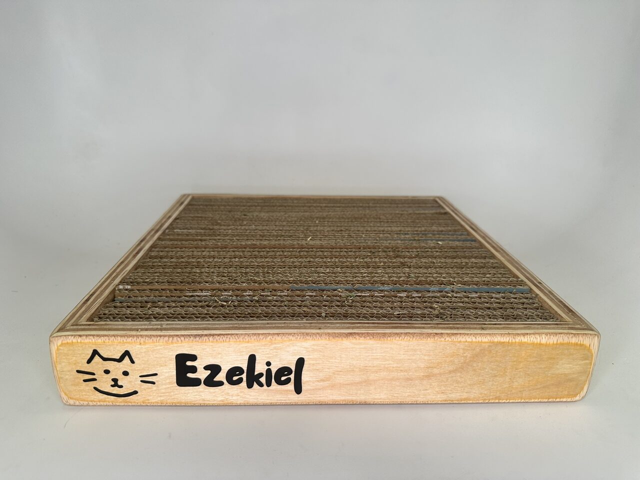 Front view of the plywood scratcher box on a white backdrop, with a black line-drawn cat face and the name Ezekiel on its front edge and corrugated cardboard filling the top.
