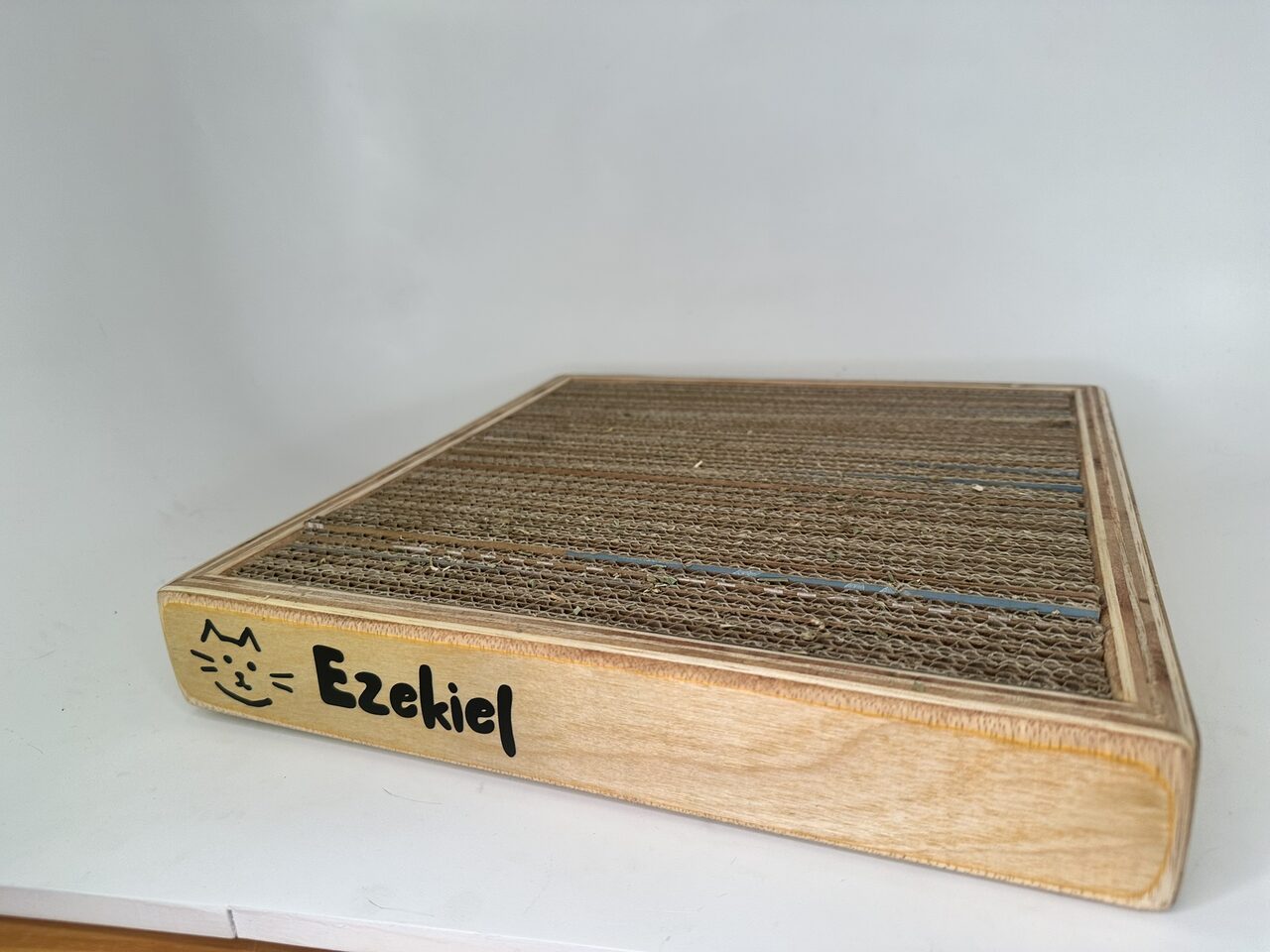 Low corner view of the scratcher box showing the plywood edge layers and the cat face and name on the side.