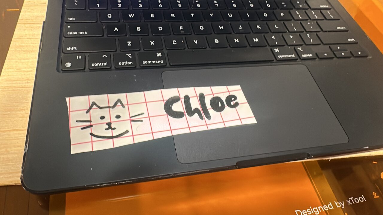 A marker drawing of a cat face and the name Chloe on red grid paper, lying on a laptop on the laser bed.