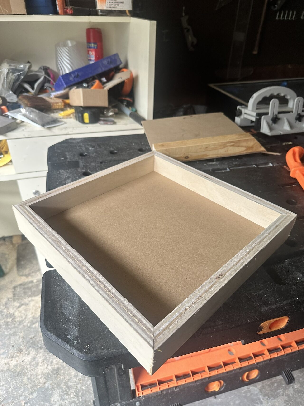 An empty shallow plywood tray with a hardboard base sitting on a work stand in the garage.