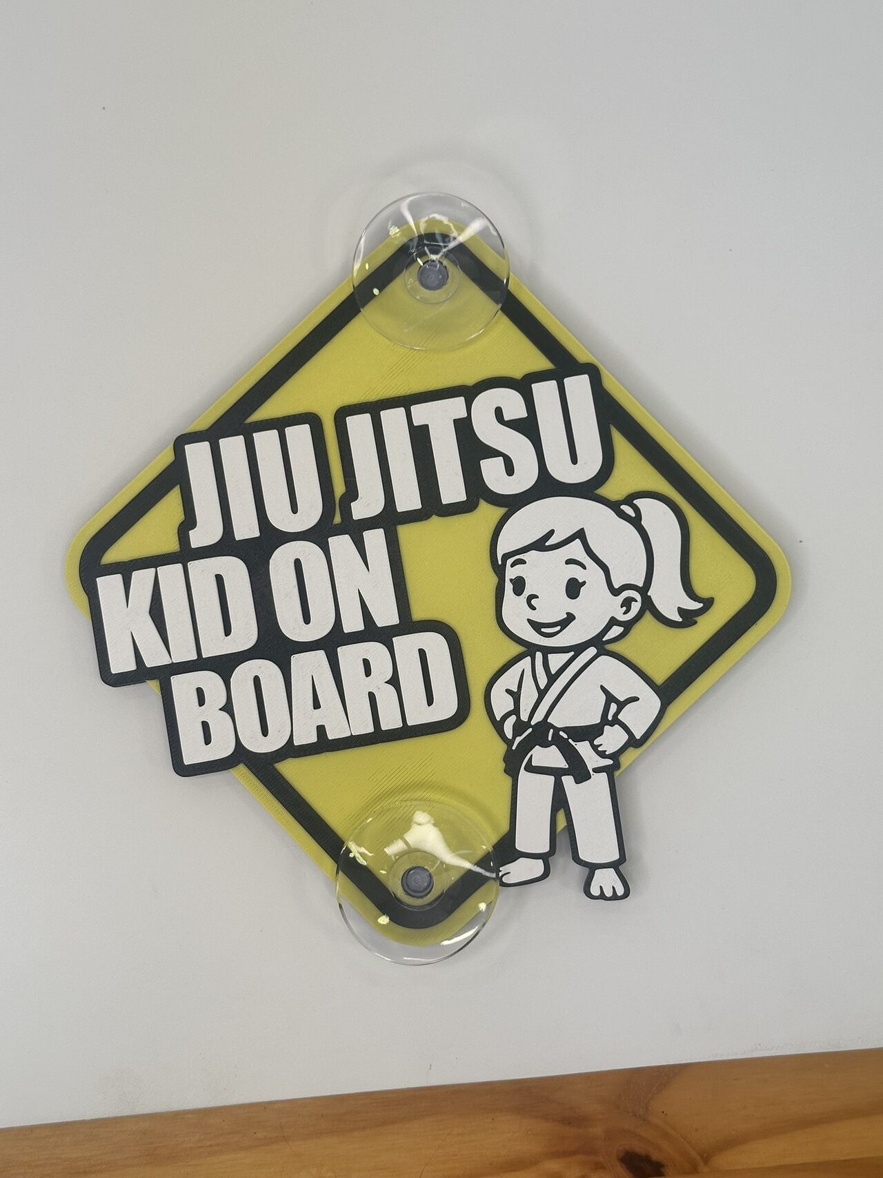 Yellow diamond sign held to a white wall by two clear suction cups, with raised white lettering outlined in black reading Jiu Jitsu Kid On Board and a cartoon girl with a ponytail in a white gi and black belt.