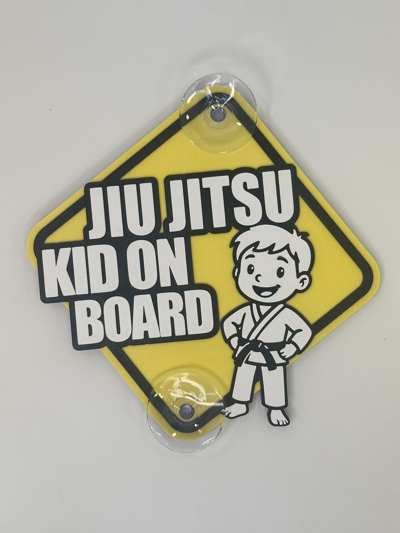 The boy version: yellow diamond sign on a white wall reading Jiu Jitsu Kid On Board with a cartoon boy in a white gi and black belt, hands on hips.