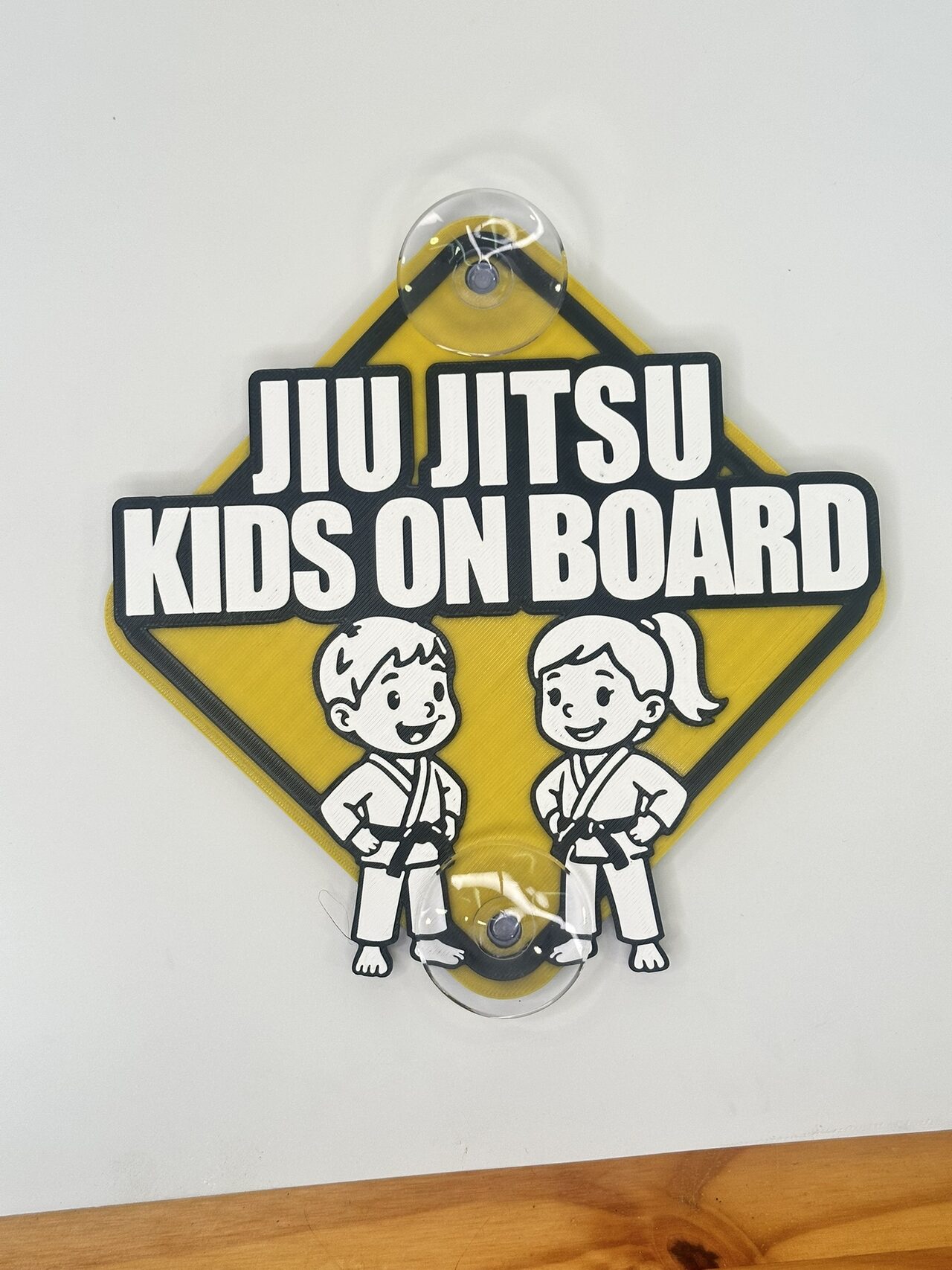 The two-kid version: yellow diamond sign on a white wall reading Jiu Jitsu Kids On Board above a cartoon boy and girl in white gis and black belts.