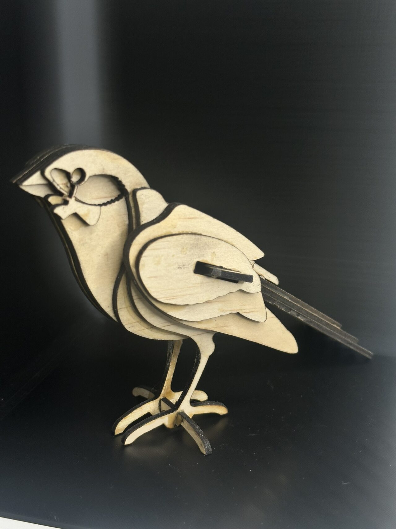 Small bird figure made of stacked flat pale wood layers with dark cut edges, standing on slotted wood feet against a black backdrop, lit from the side.