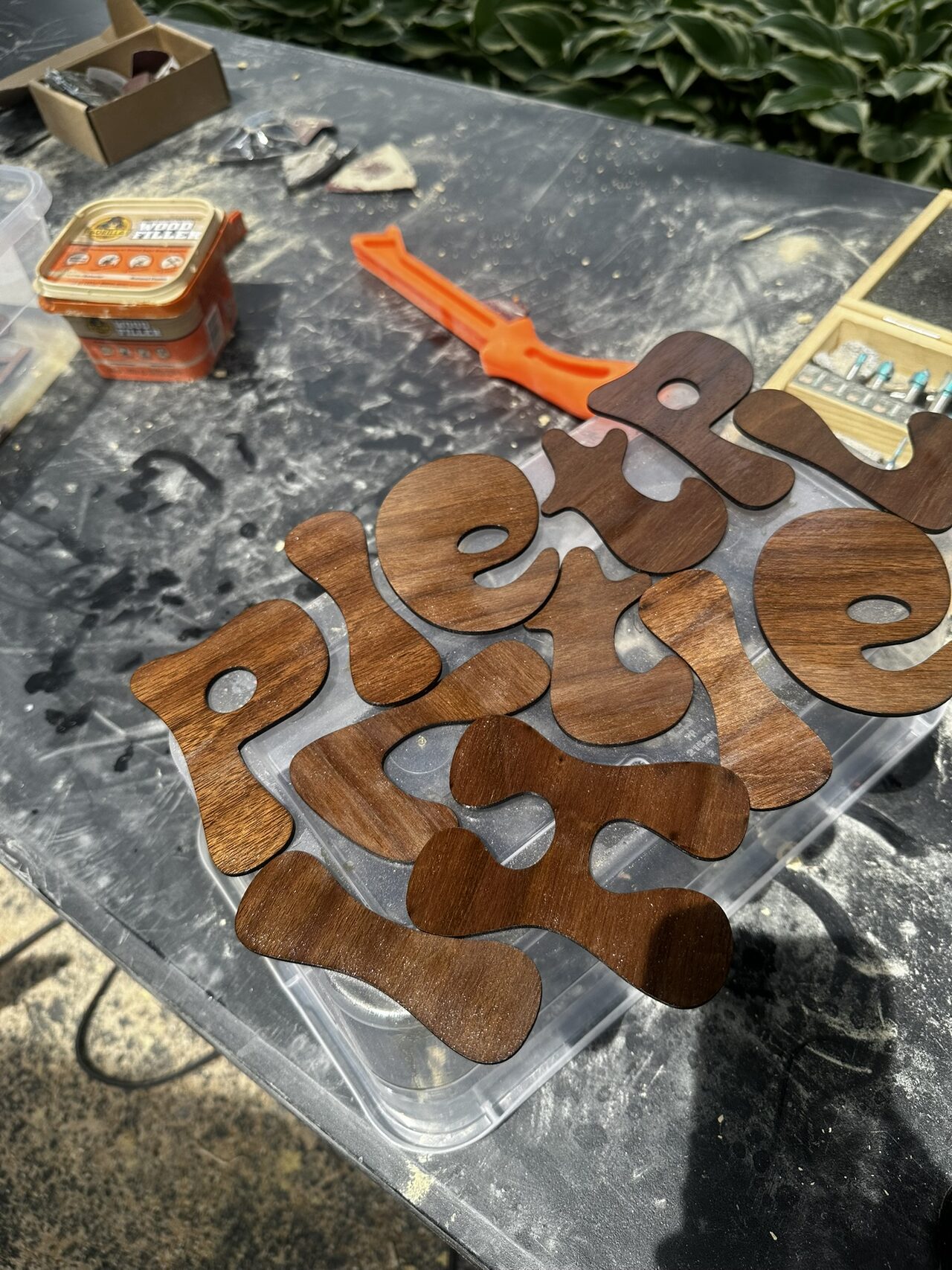 Laser cut bubble letters stained a rich dark brown, laid out to dry on a clear plastic sheet on a dusty work table.
