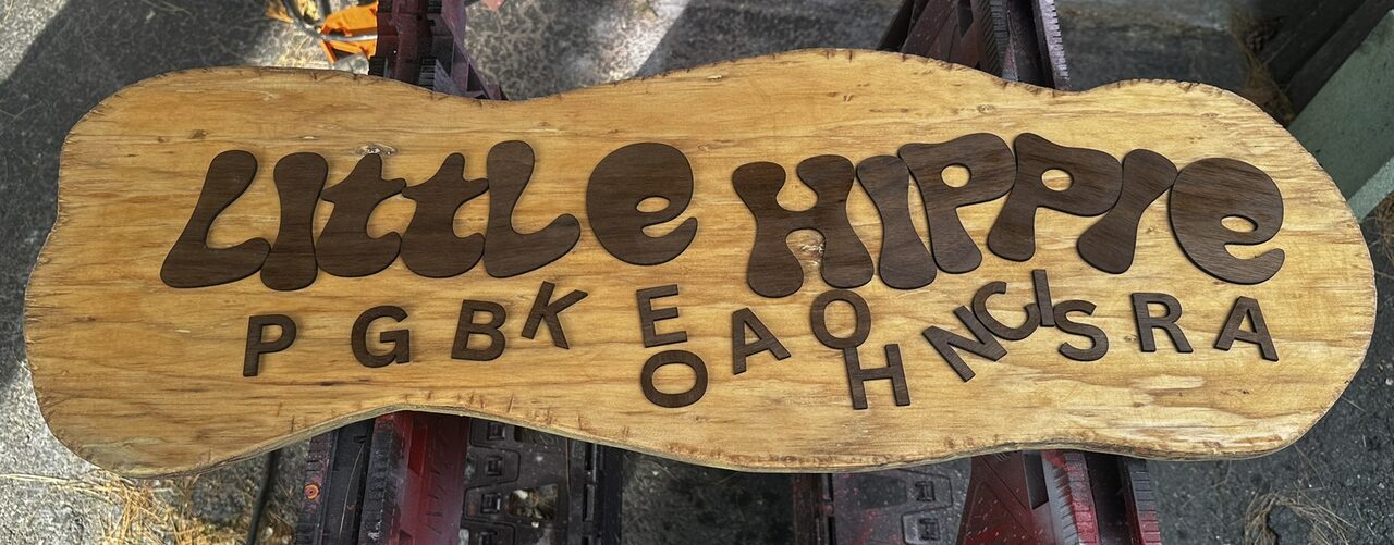 A golden stained plywood board with a wavy outline on sawhorses, with dark raised letters reading Little Hippie and the smaller letters of the second line laid out below.