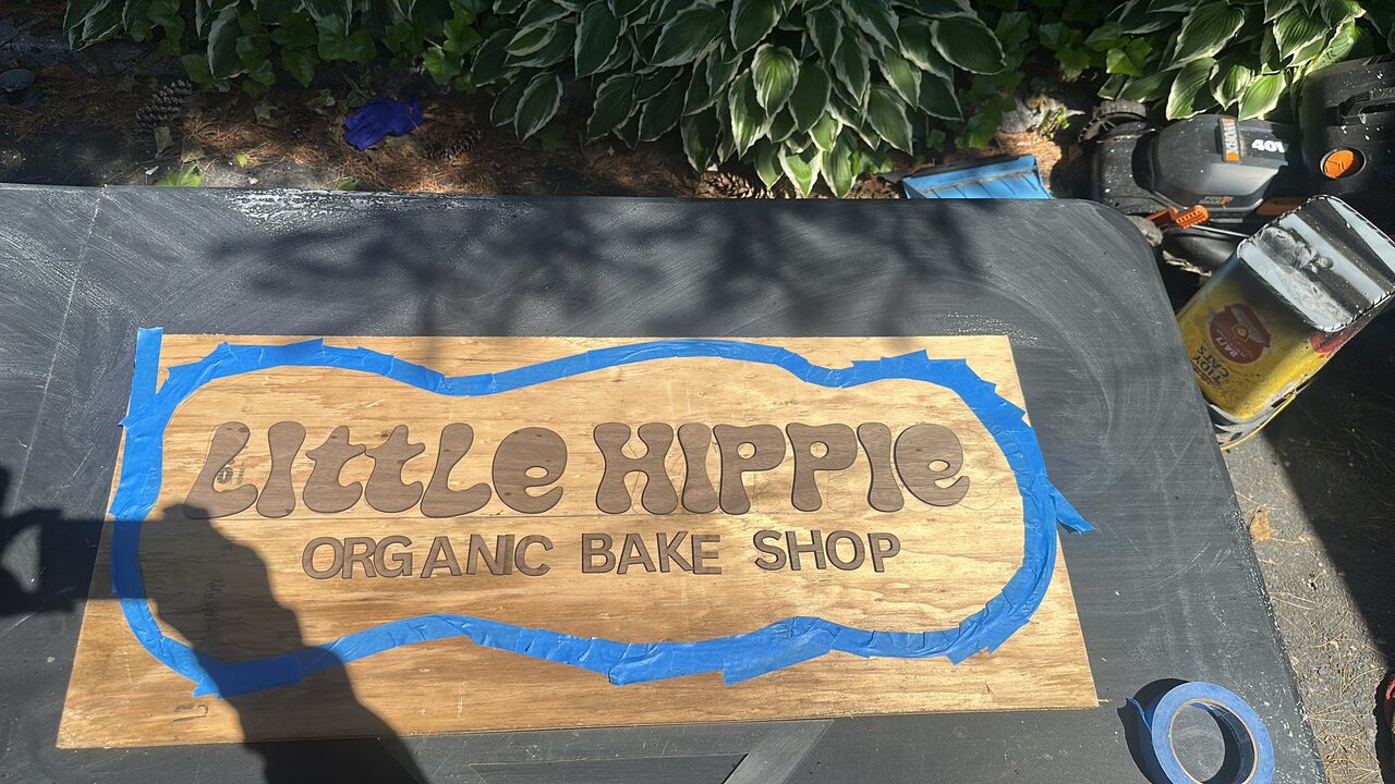 The sign's lettering laid out on a plywood sheet on a work table, with blue tape marking a wavy outline around it.
