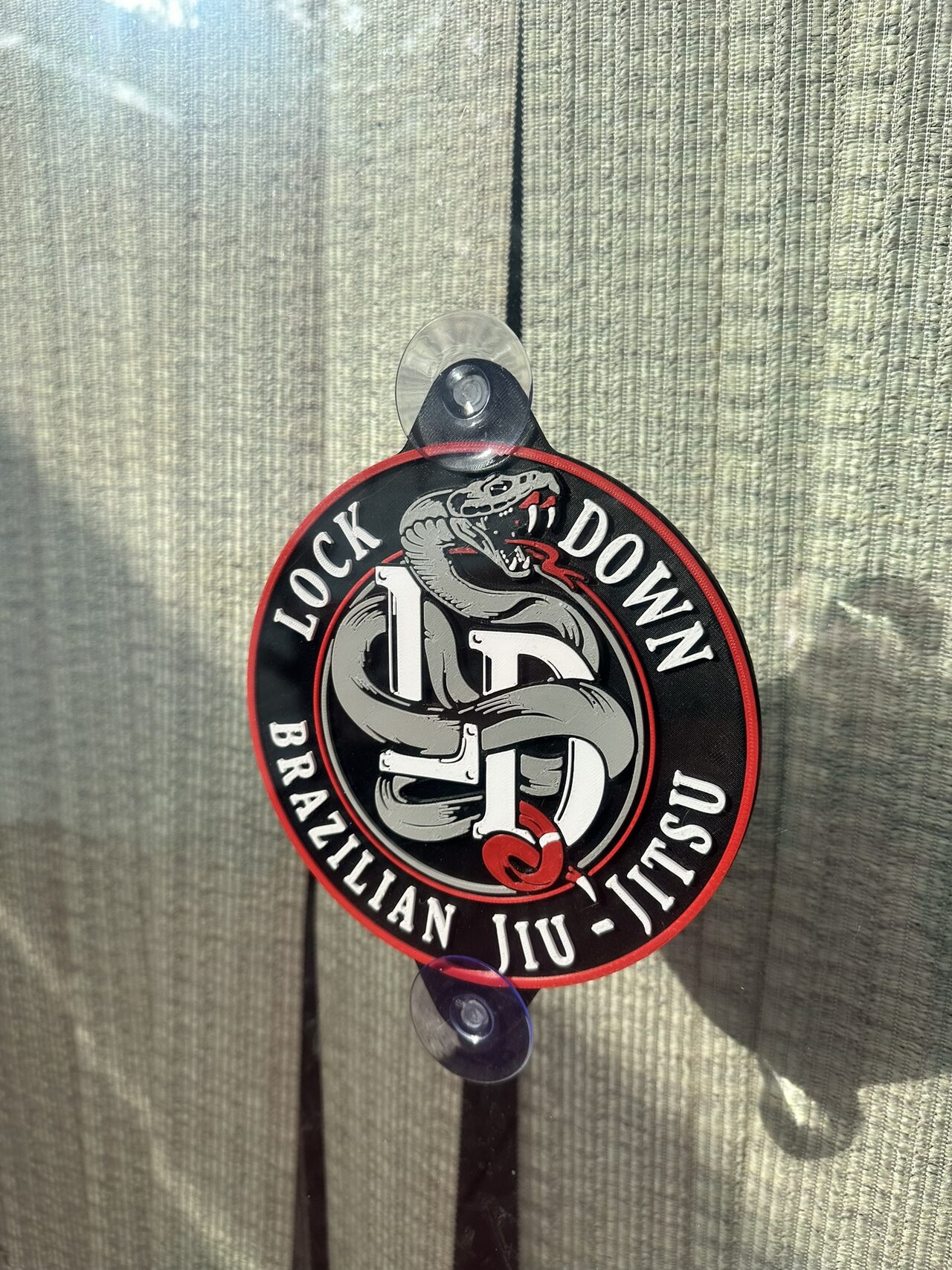 Round logo plaque hung on a window by two suction cups in front of closed blinds: a grey snake coiled through a white LD monogram, ringed by raised white lettering reading Lock Down Brazilian Jiu-Jitsu, with a red rim.