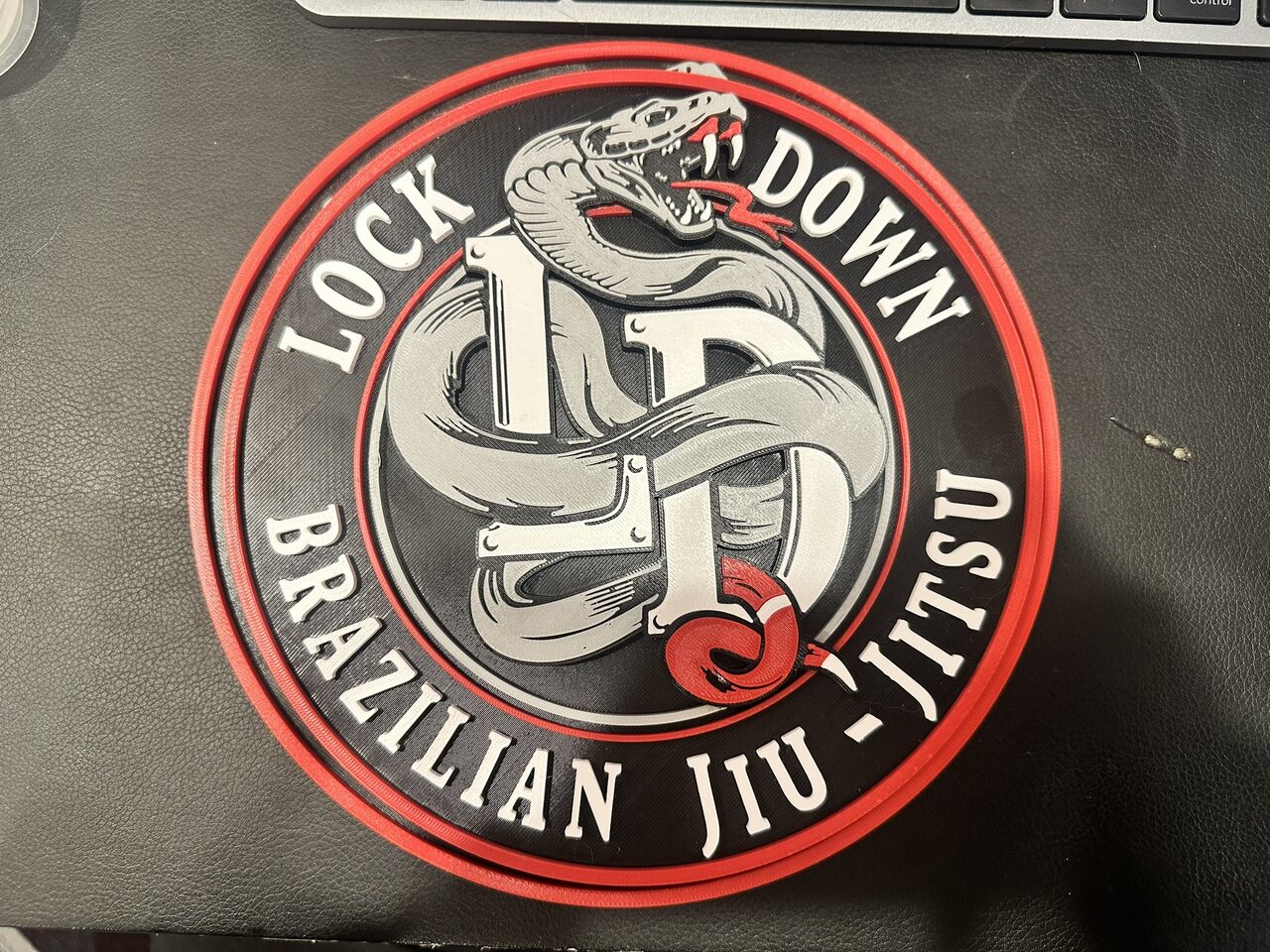The round Lock Down Brazilian Jiu-Jitsu plaque seen from above on a black desk mat, all lettering readable, a keyboard at the top edge.