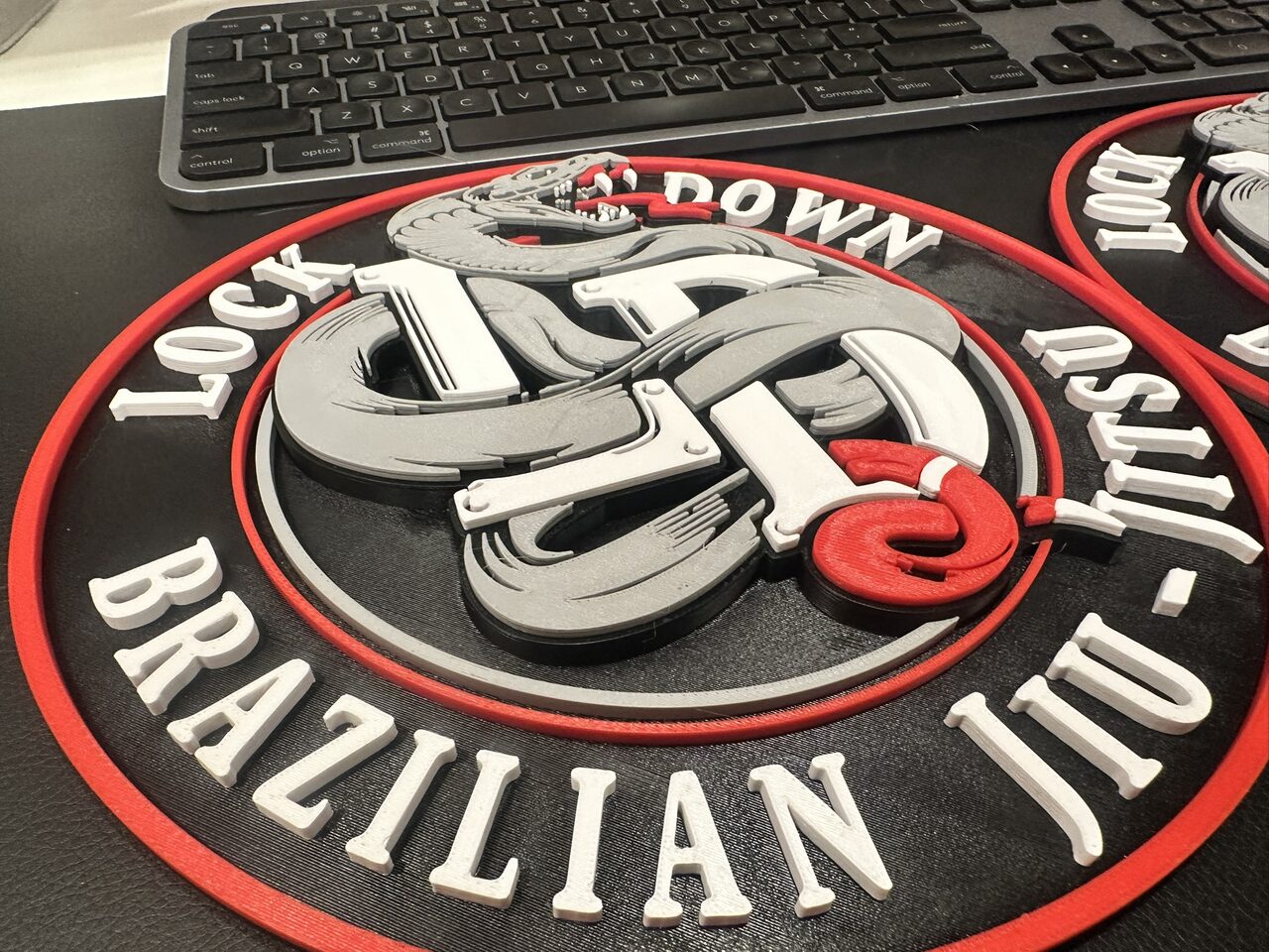 Close angled view of the plaque showing the snake, monogram and letters standing up from the black background in separate raised layers.