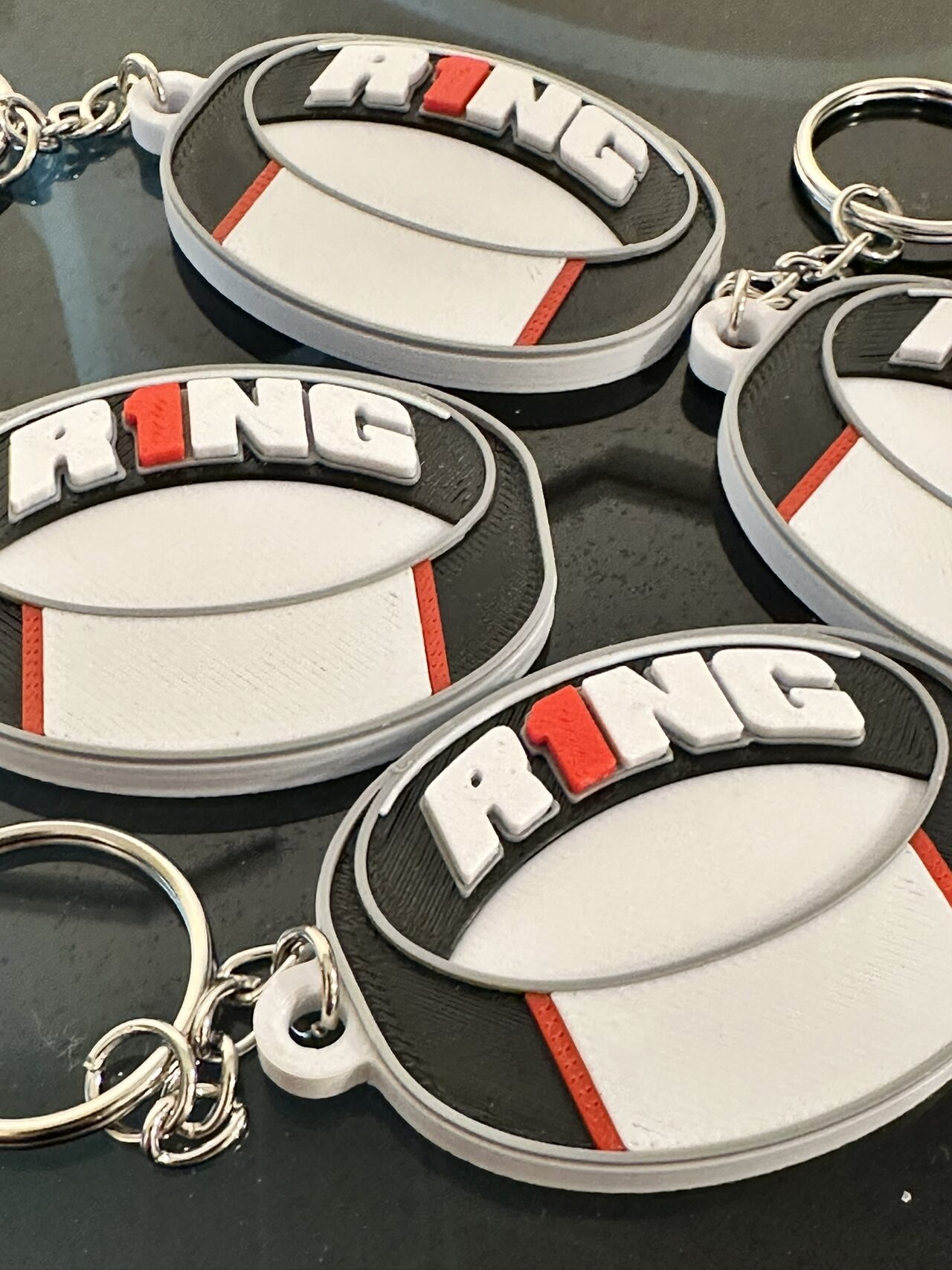 Close-up of several oval R1NG keychains printed in white, black and red, each with a chain and split ring, lying on a dark surface.