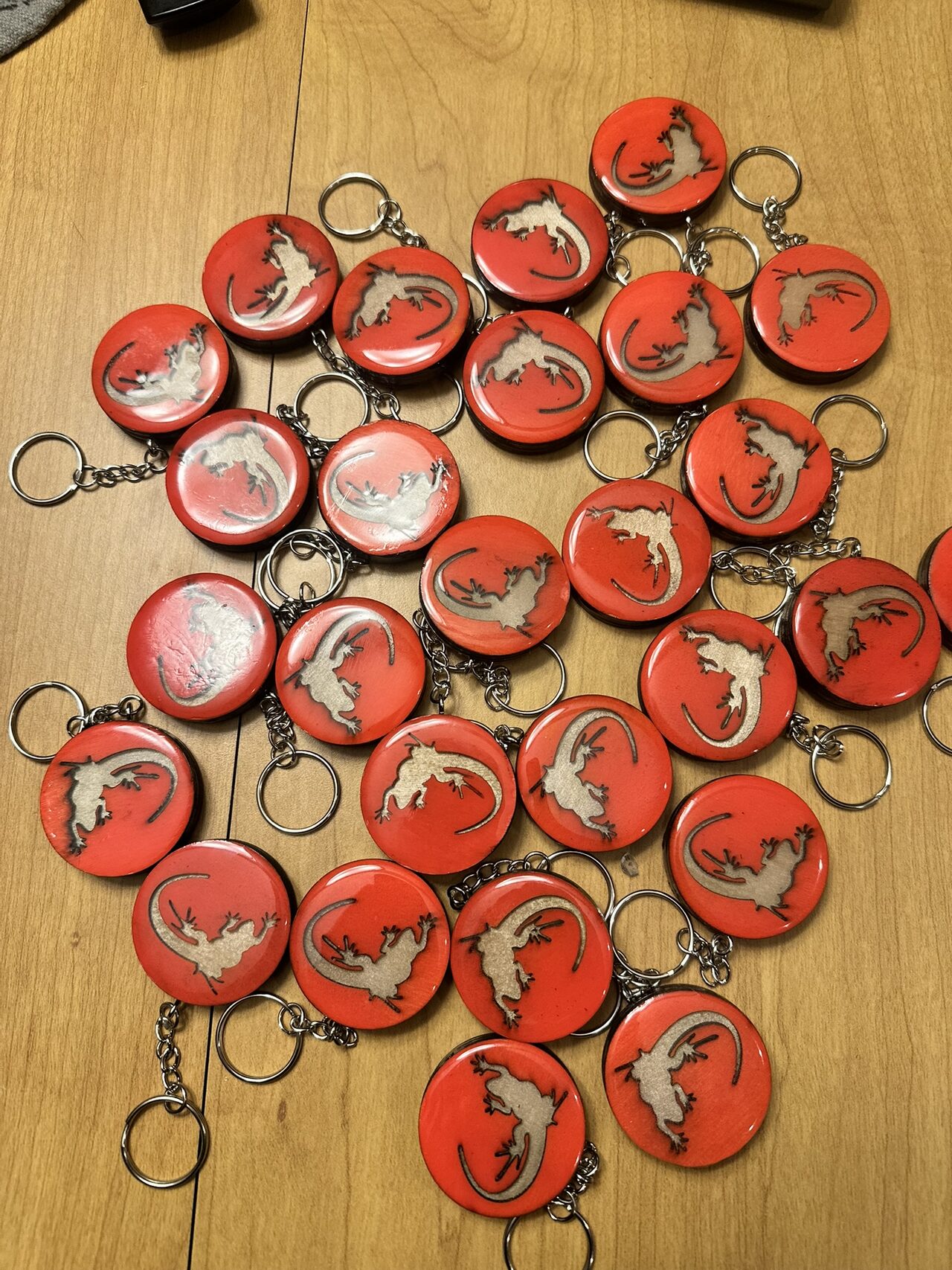 About thirty round red keychains with a pale lizard inlay and a glossy domed top, with chains and rings, spread on a wood table and seen from above.