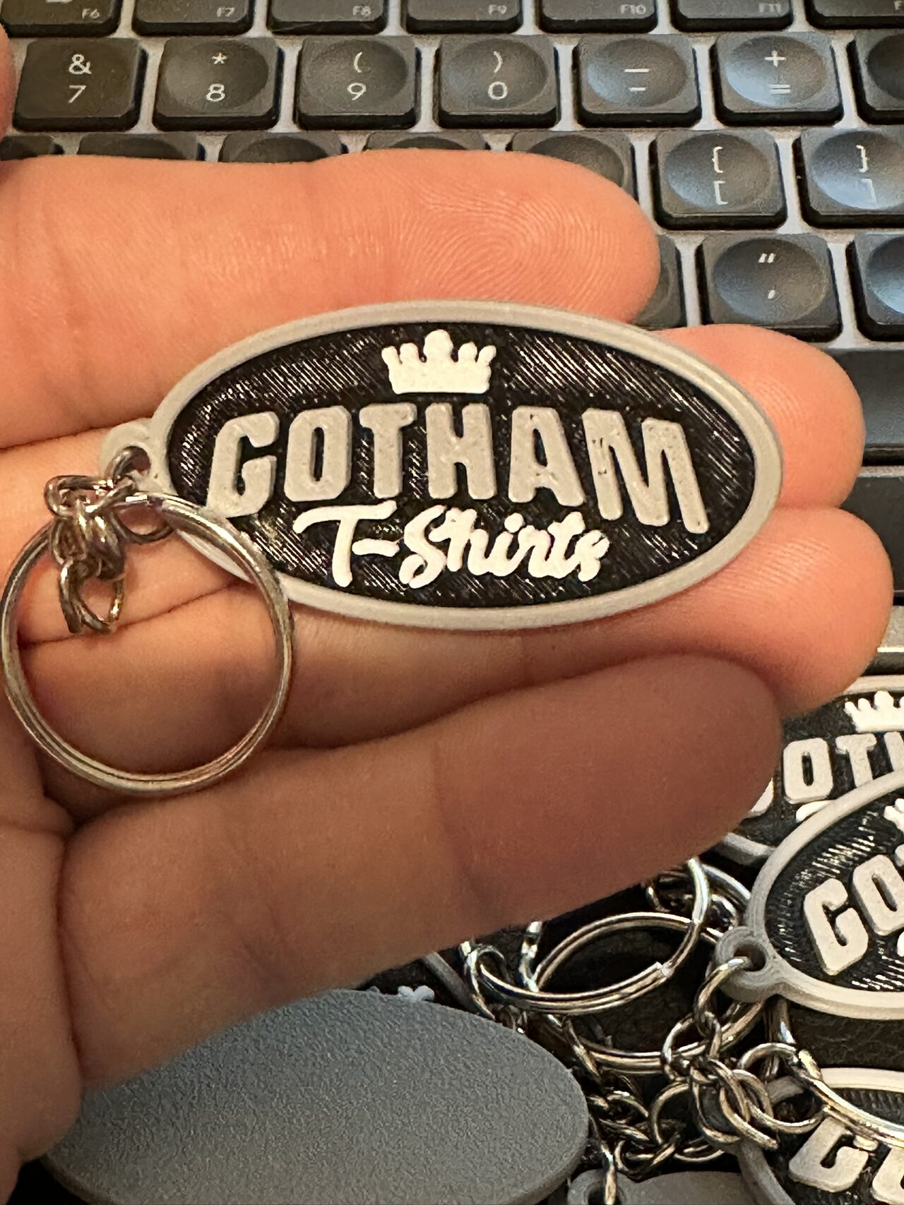An oval black and white Gotham T-Shirts keychain with a crown above the lettering, held in a palm with a pile of the same keychains behind.