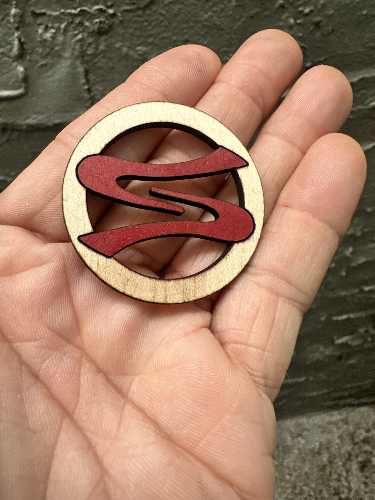 A round wood keychain disc with a dark red S logo across an open ring, held in an open palm.
