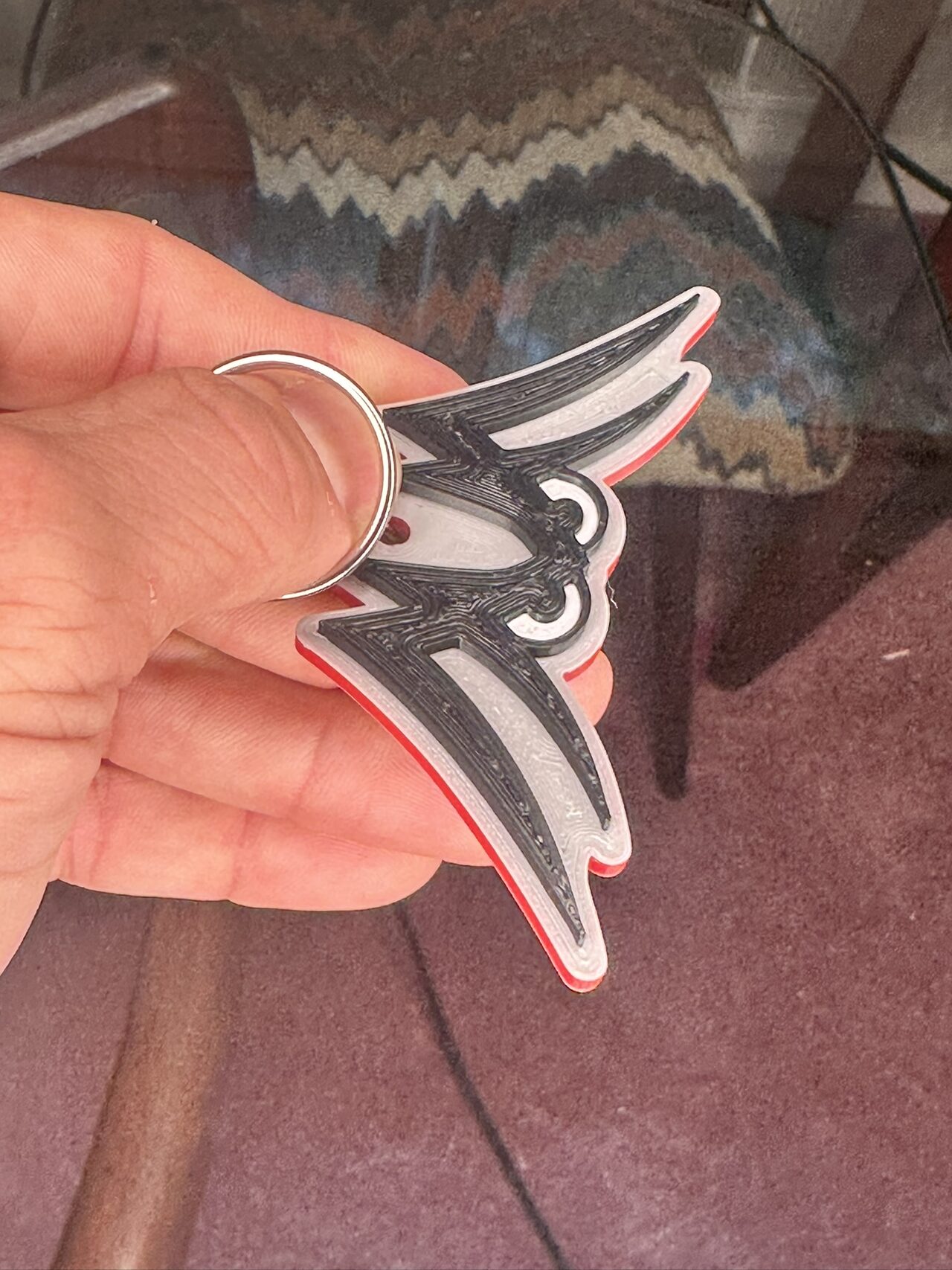 A winged-eyes logo keychain printed in black and silver-white with a red back layer, held between the fingers by its split ring.