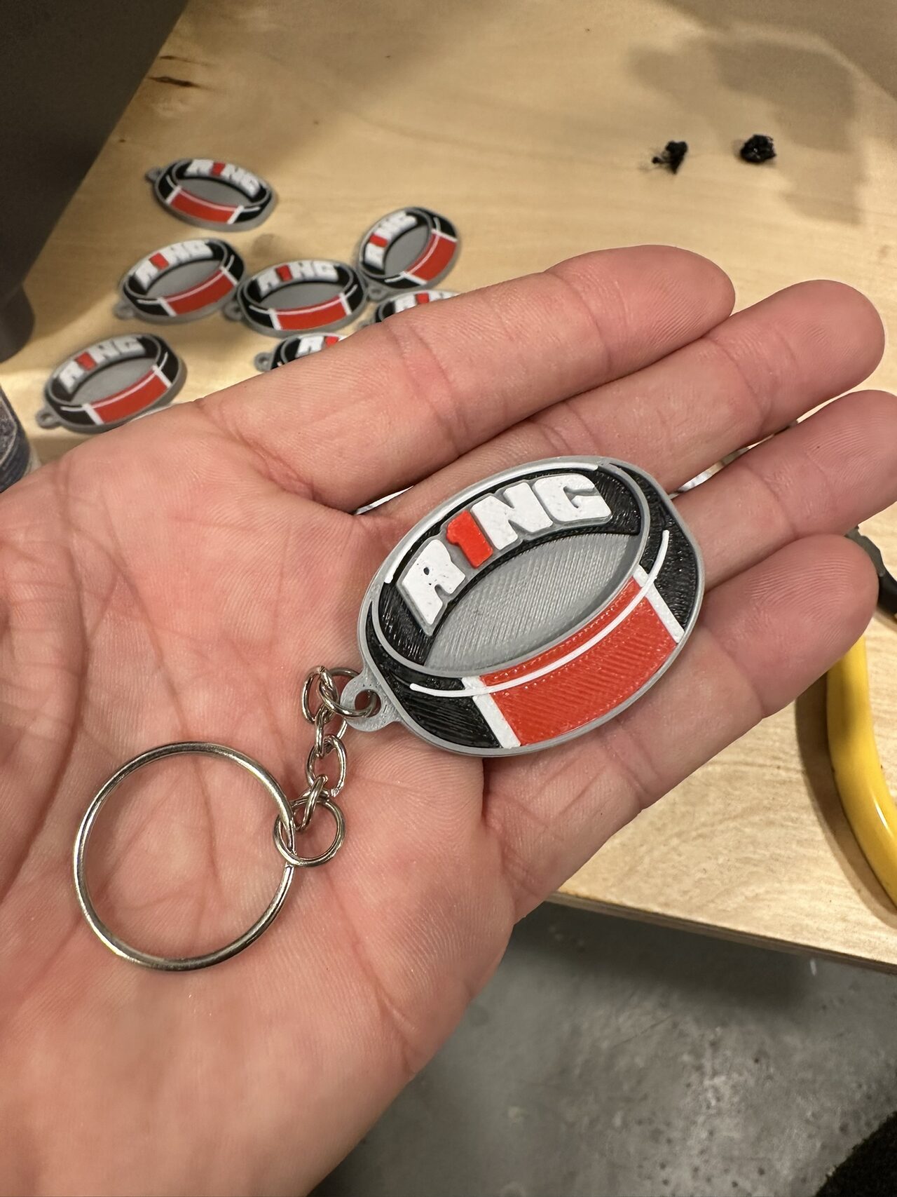 A round R1NG keychain in black, white, grey and red with a chain and split ring in an open palm, more keychains on the table behind.