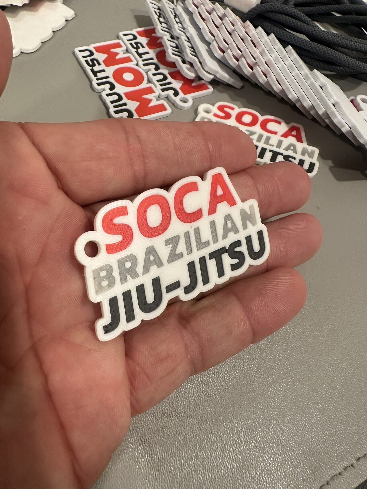 A keychain cut to the shape of the words Soca Brazilian Jiu-Jitsu in red, grey and black on white, held in a palm with more lettered keychains behind.