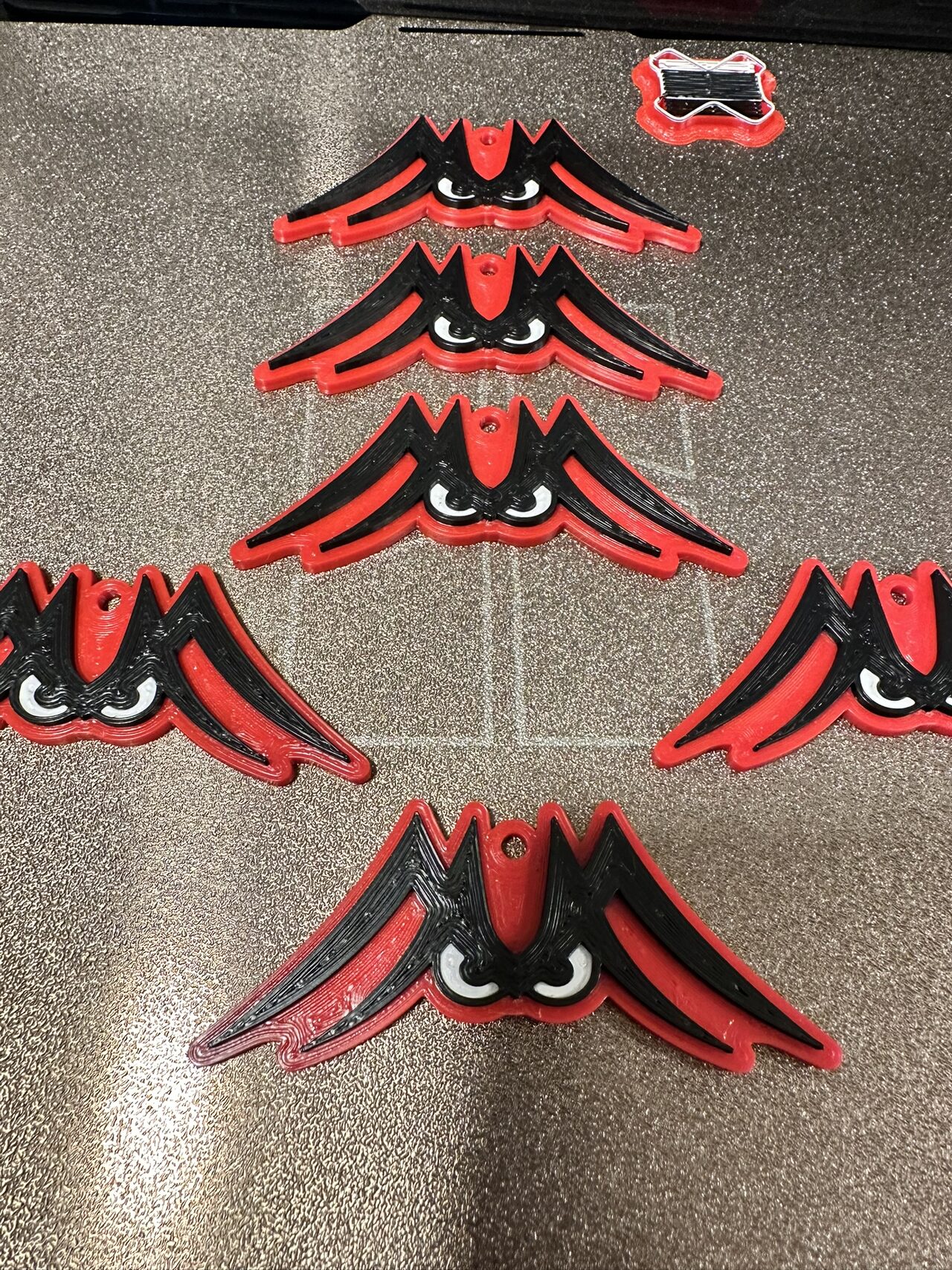 Six red and black winged-eyes keychains lying on the textured build plate of a 3D printer, seen from above.