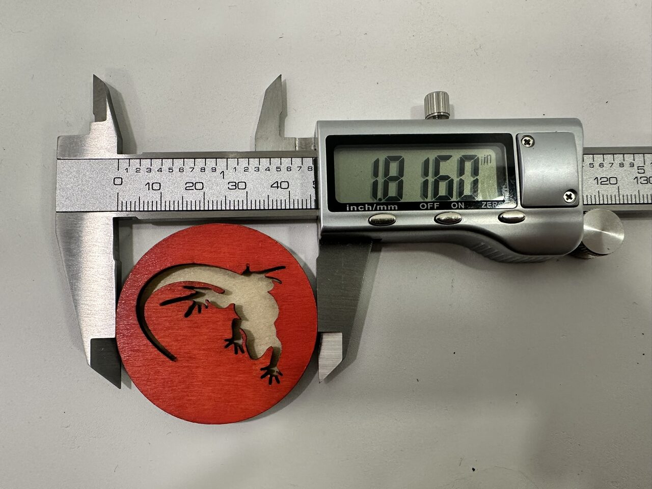 Digital calipers measuring the thickness of a red wood disc with a lizard-shaped cutout, on a white surface.