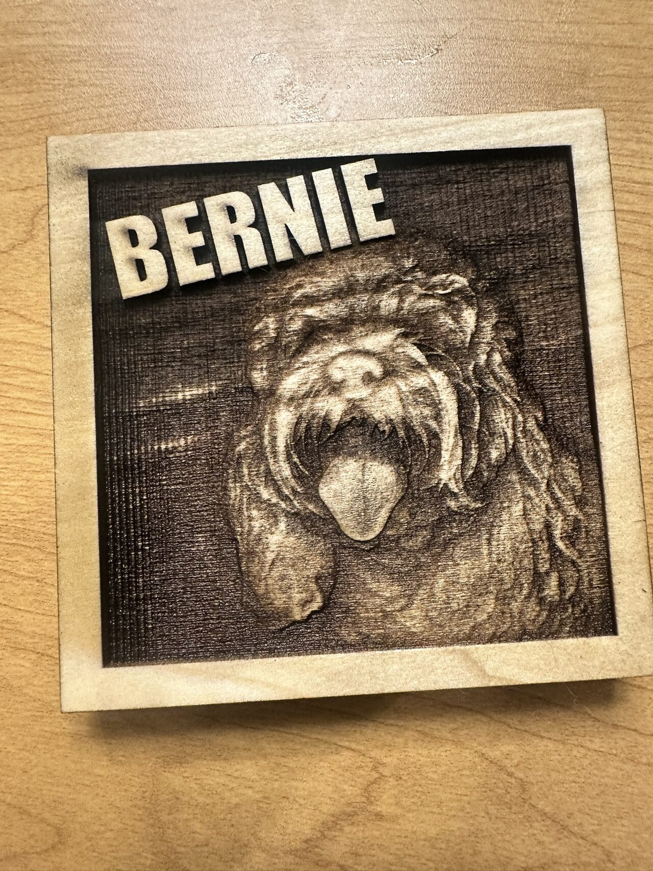 Square wood plaque engraved in relief with a shaggy dog, tongue out, and the name BERNIE in raised letters at the top left, lying on a wood table.
