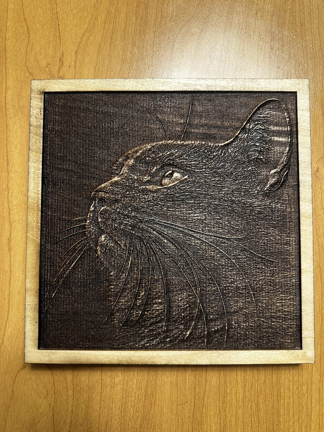 Square wood plaque engraved with a cat's head in profile looking up, long whiskers cut as fine lines, pale border.