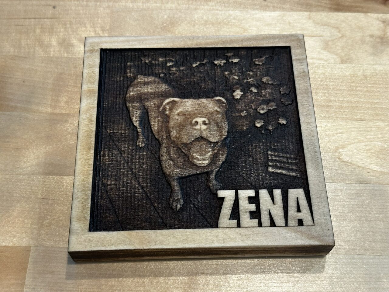 Wood plaque engraved with a smiling dog seen from above among fallen leaves, the name ZENA at bottom right.