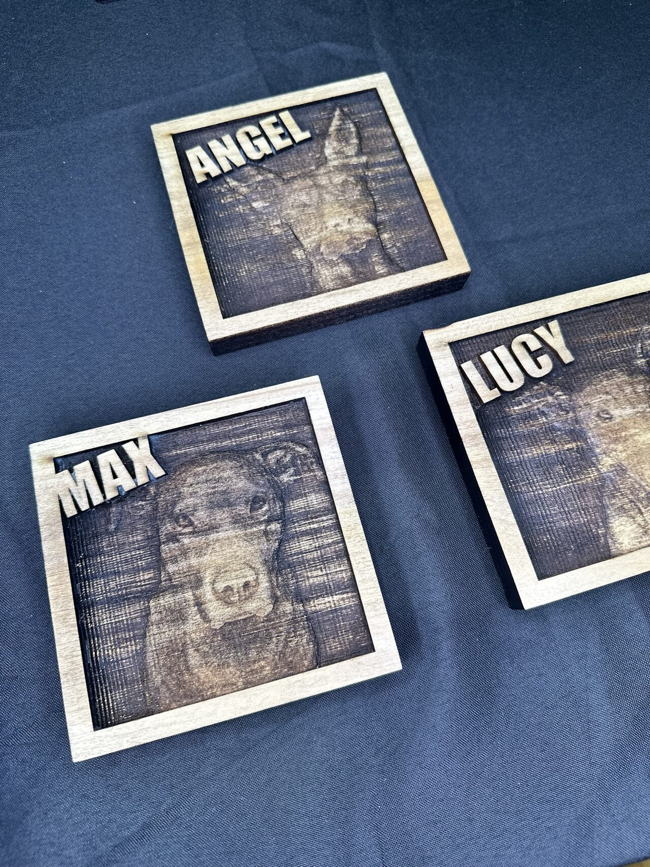 Three square dog portrait plaques named ANGEL, MAX and LUCY laid out on a dark blue cloth.