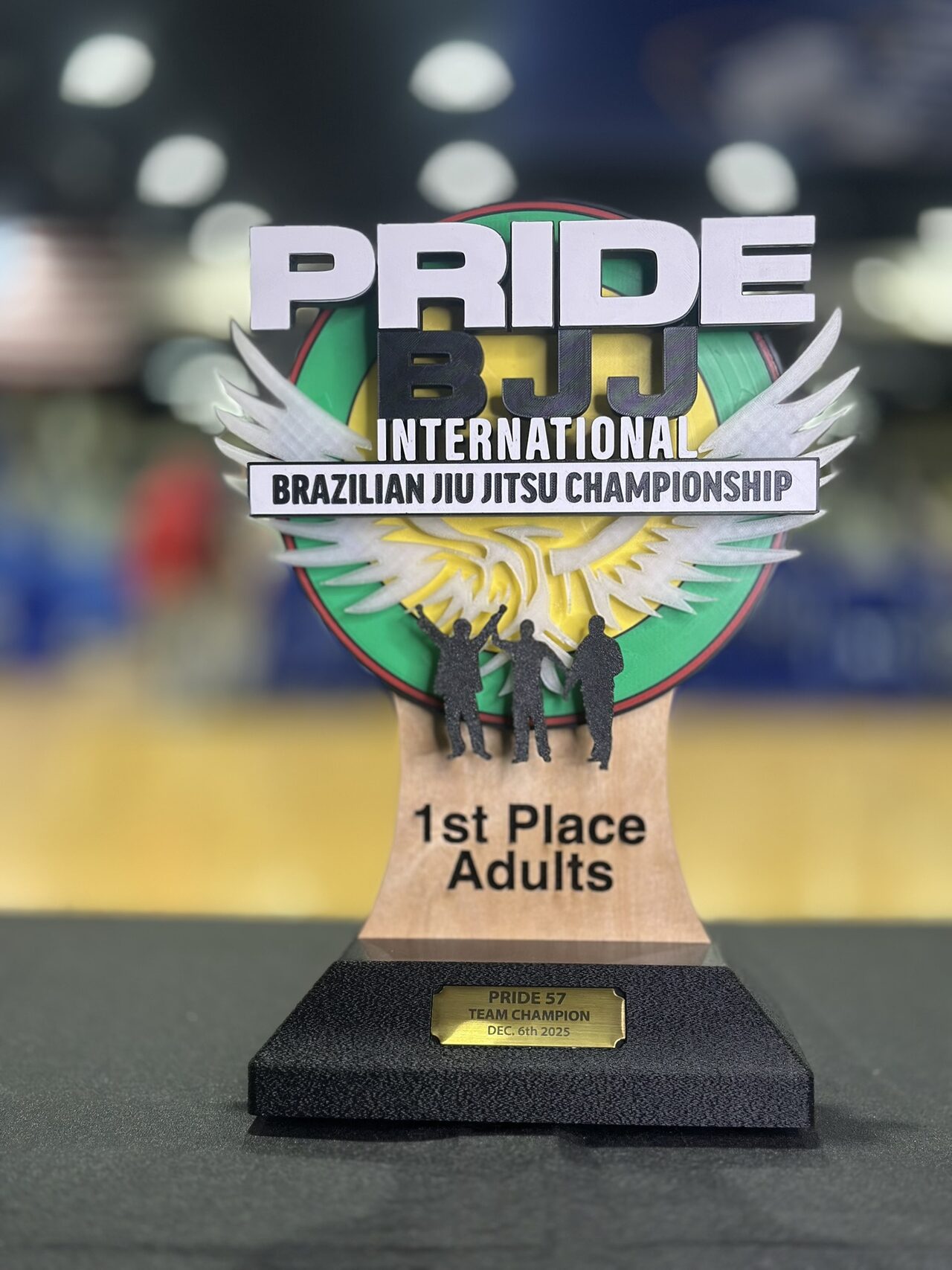 A trophy on a black tablecloth: raised white PRIDE and black BJJ lettering over a green and yellow disc with white wings and three black figures, on a wood body engraved 1st Place Adults and a black base with a gold-coloured plate.