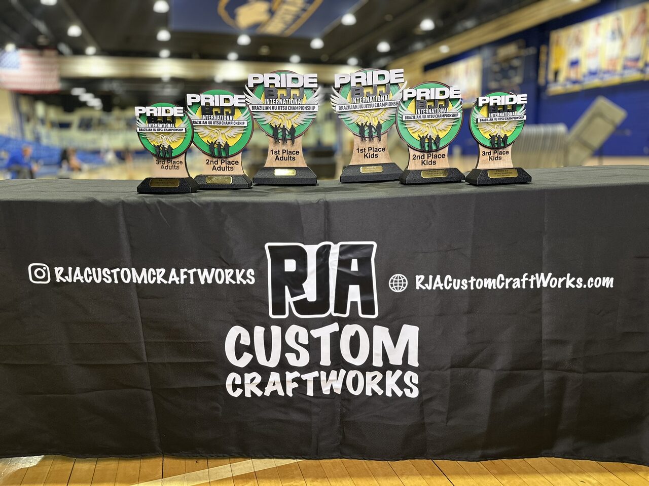The six trophies lined up on a table with a black RJA Custom Craftworks tablecloth in a sports hall.