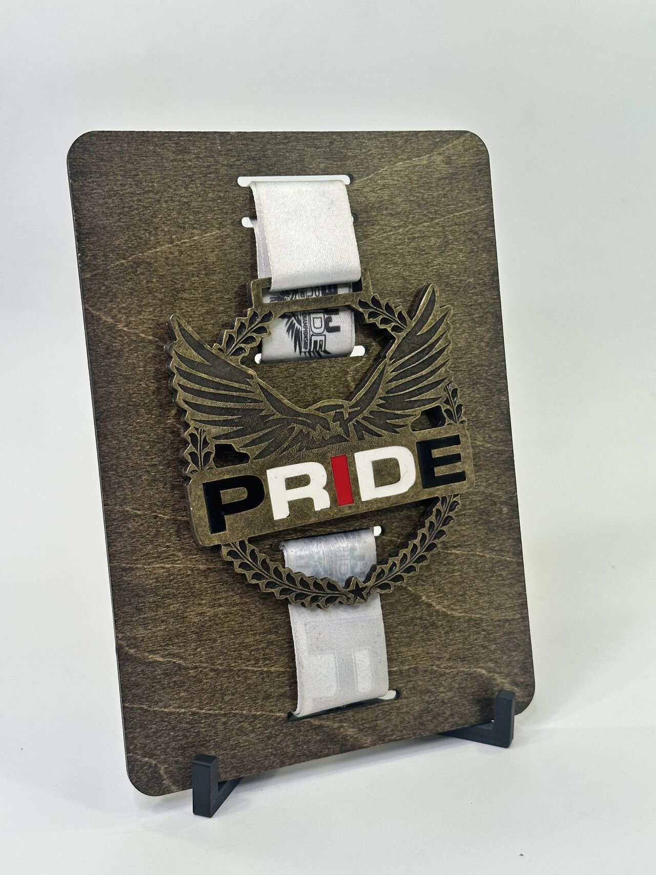 Dark stained wood board on a small black stand, holding a bronze-coloured eagle medal lettered PRIDE by a white ribbon threaded through slots above and below it, on a white backdrop.