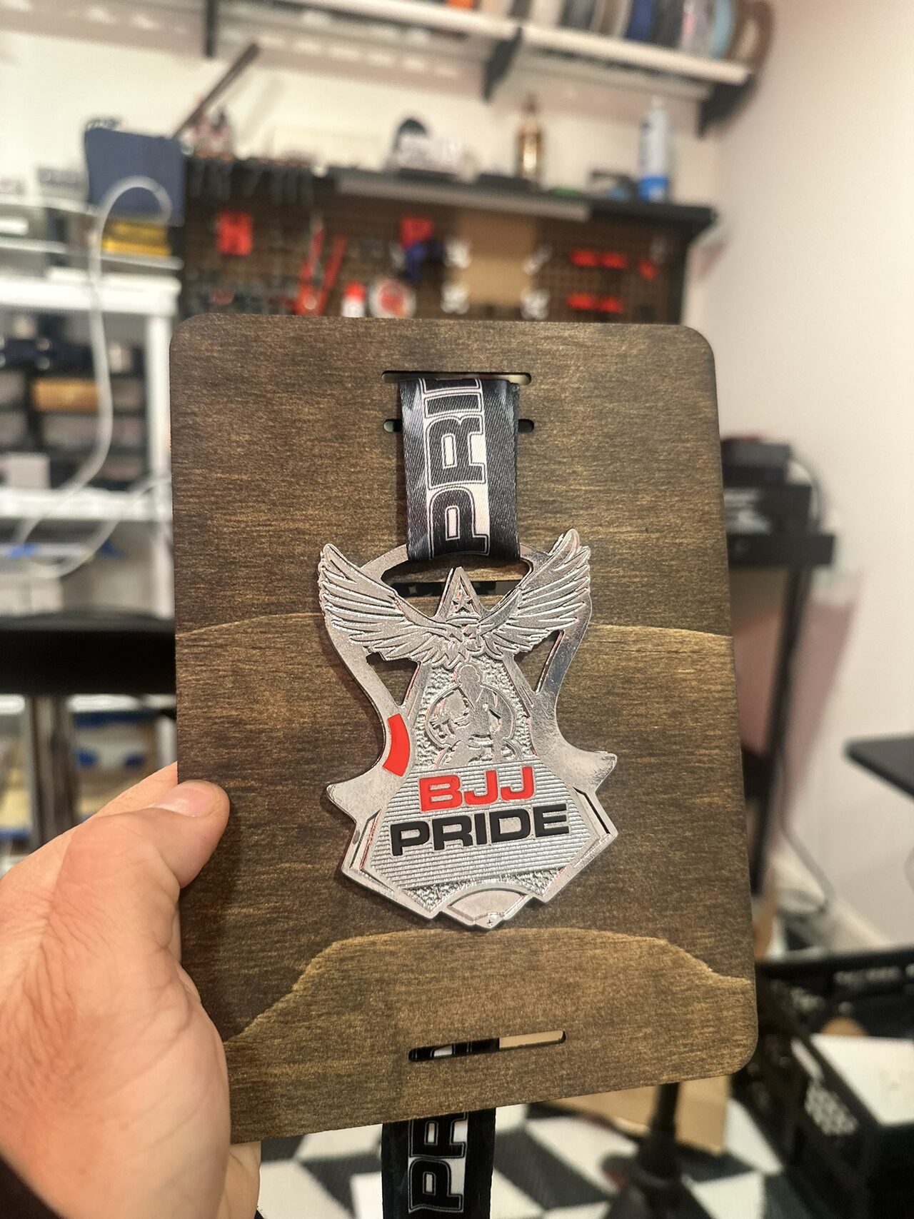 A hand holding an early stained wood board with a silver BJJ PRIDE eagle medal on a black and white ribbon, shop tool wall behind.