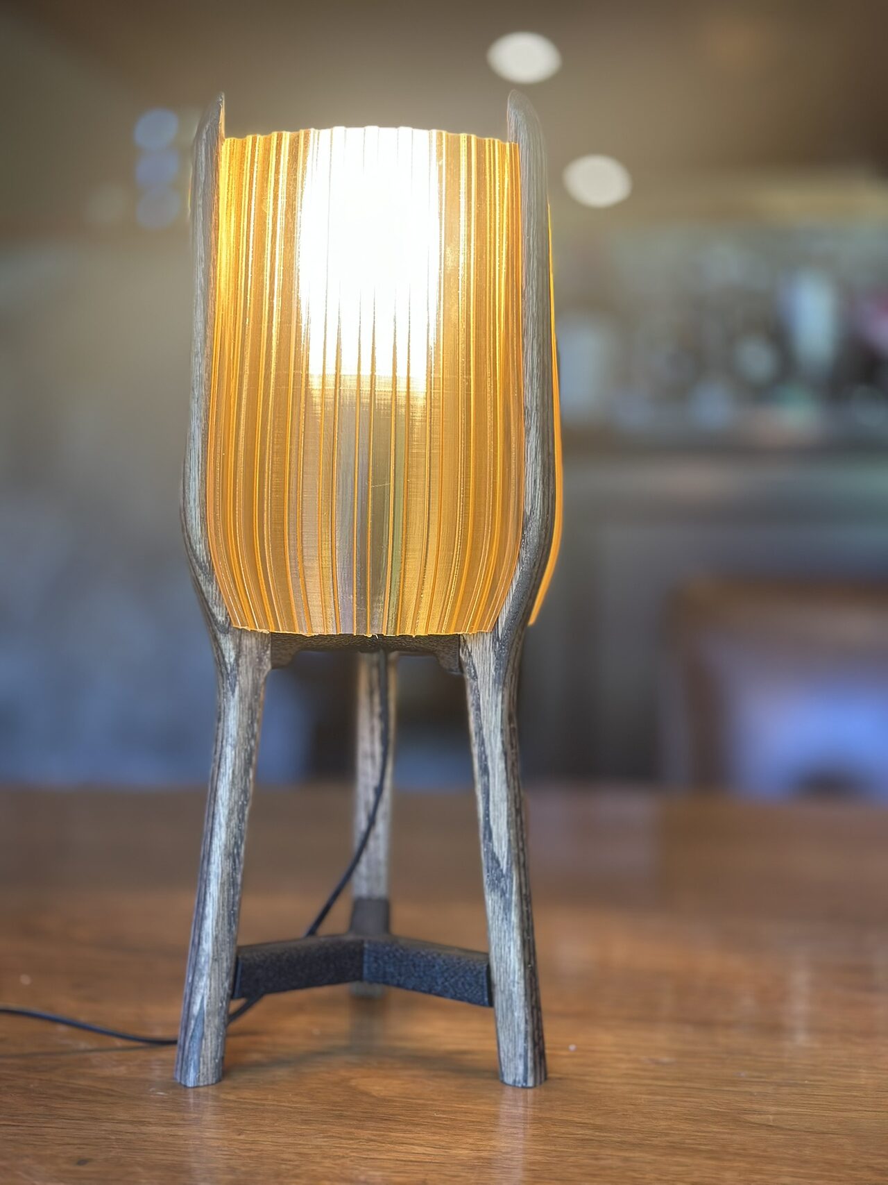 Table lamp switched on, its ribbed amber shade glowing between three grey-brown wooden legs joined by a black brace, on a wooden table with a blurred room behind