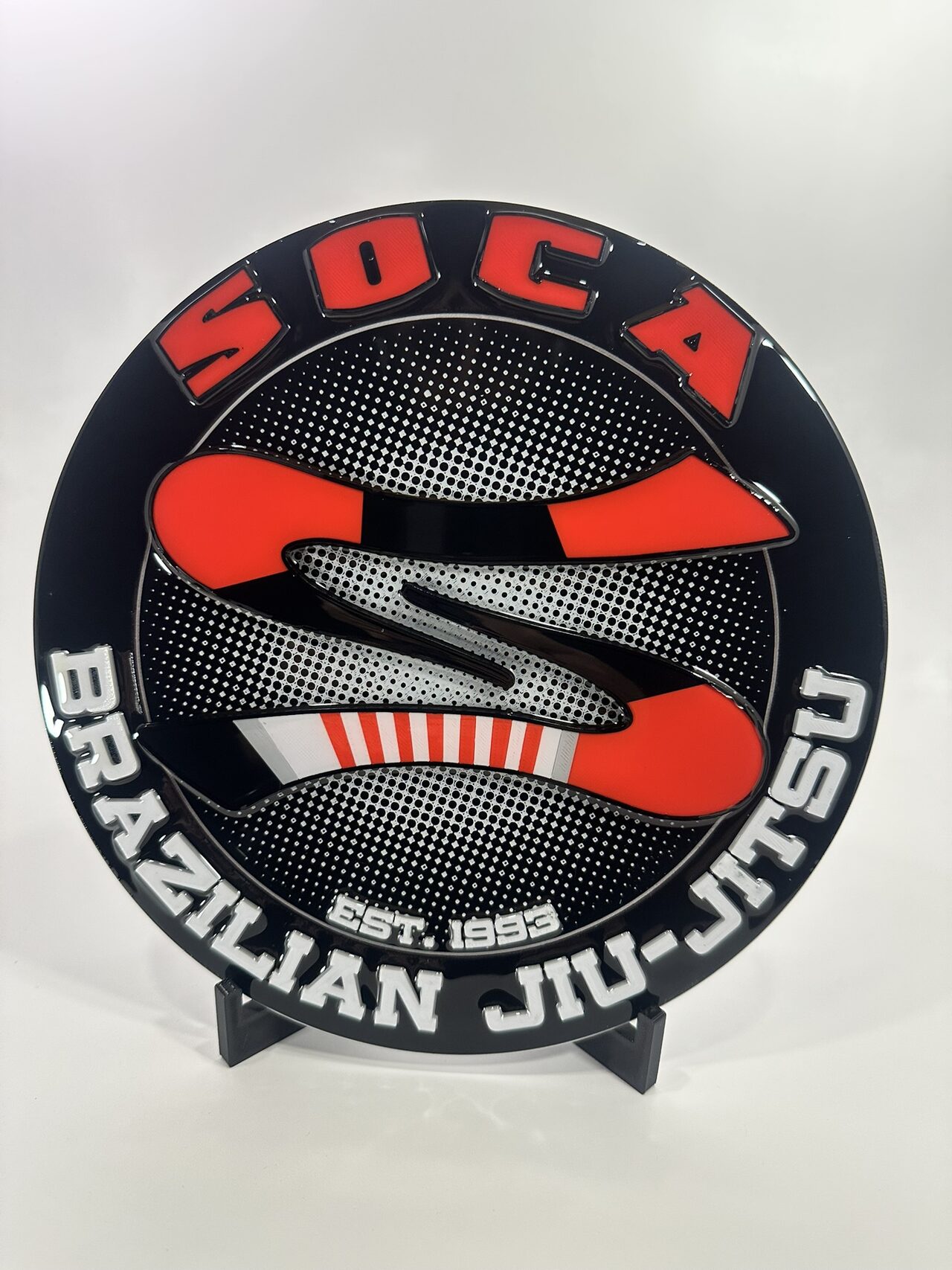 Round glossy medallion with red SOCA lettering, a red and black S-shaped belt and white Brazilian Jiu-Jitsu lettering, standing on a black easel against a white backdrop