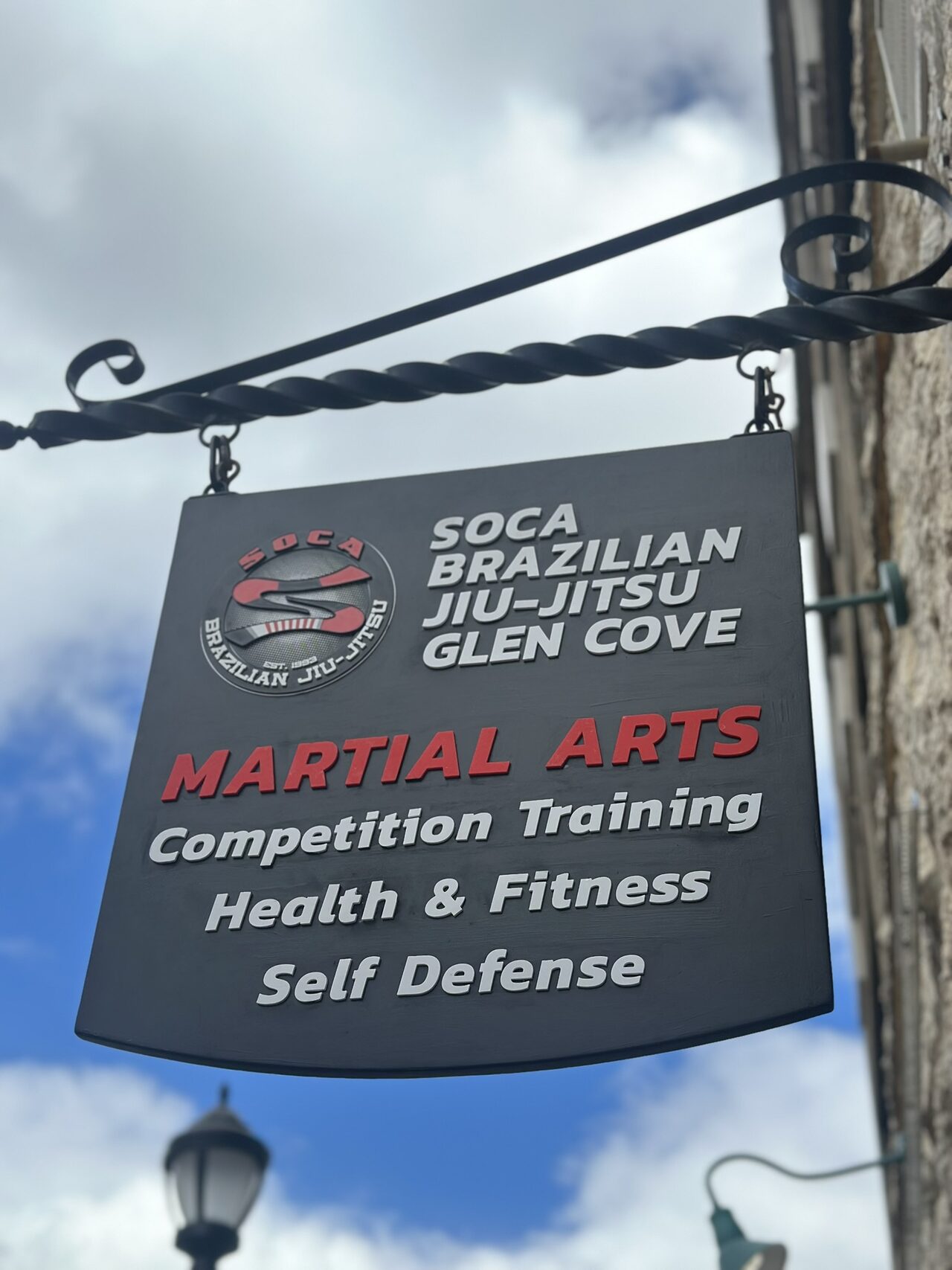 Black hanging sign on a twisted iron bracket against blue sky and clouds, with a round Soca logo and raised lettering: Soca Brazilian Jiu-Jitsu Glen Cove, Martial Arts, Competition Training, Health & Fitness, Self Defense.