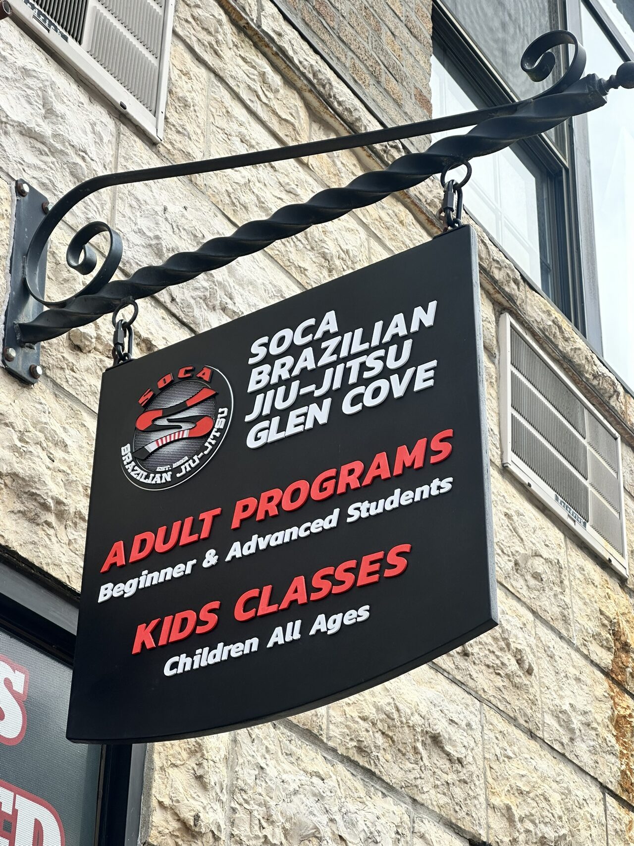 Hanging black sign on an iron bracket beside a stone wall, reading Soca Brazilian Jiu-Jitsu Glen Cove, Adult Programs, Beginner & Advanced Students, Kids Classes, Children All Ages.