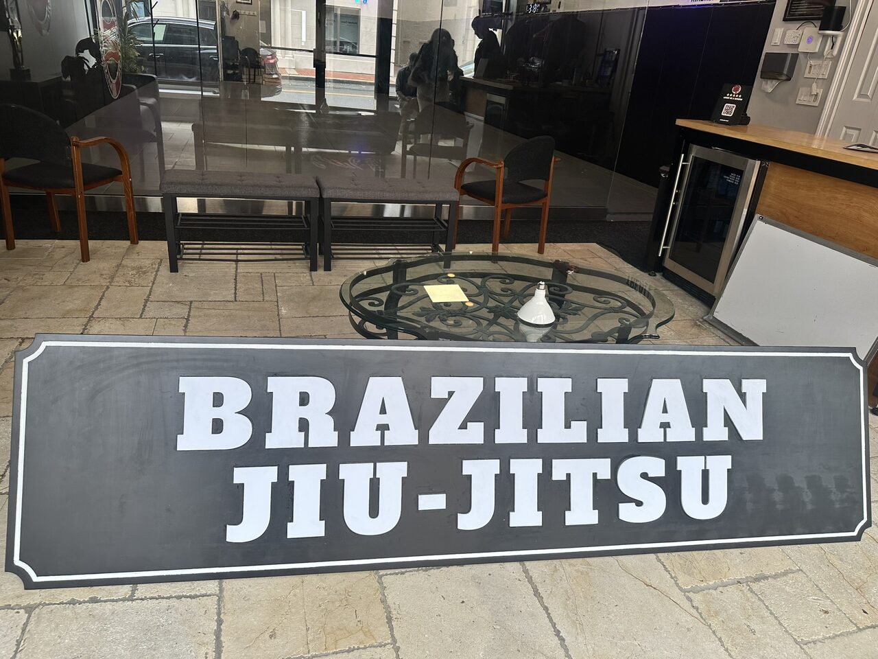 Long black panel with a white pinstripe border and raised white letters reading Brazilian Jiu-Jitsu, leaning against a glass wall on a tile floor.