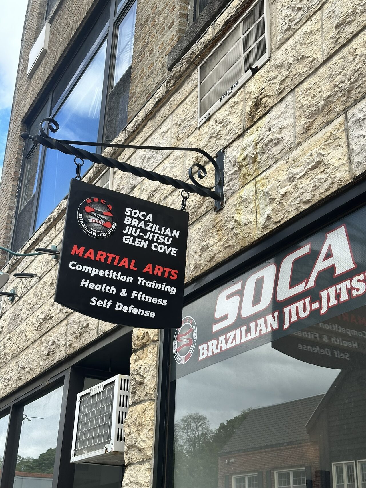 Hanging blade sign on the stone facade above a storefront window with Soca Brazilian Jiu-Jitsu window lettering.