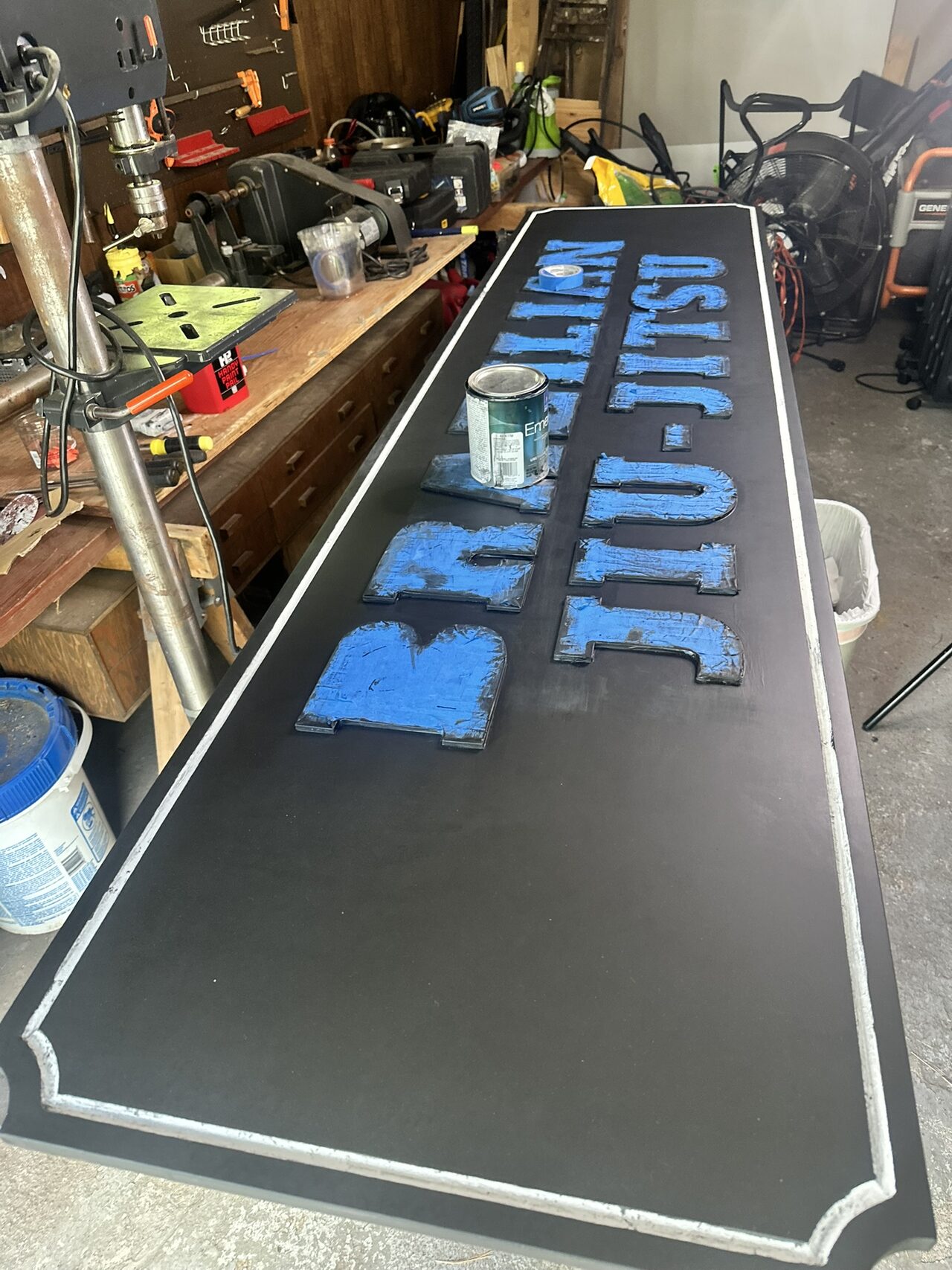 Long black panel in the workshop with its letters wrapped in blue masking tape, a freshly painted white pinstripe border and a paint can on the panel.