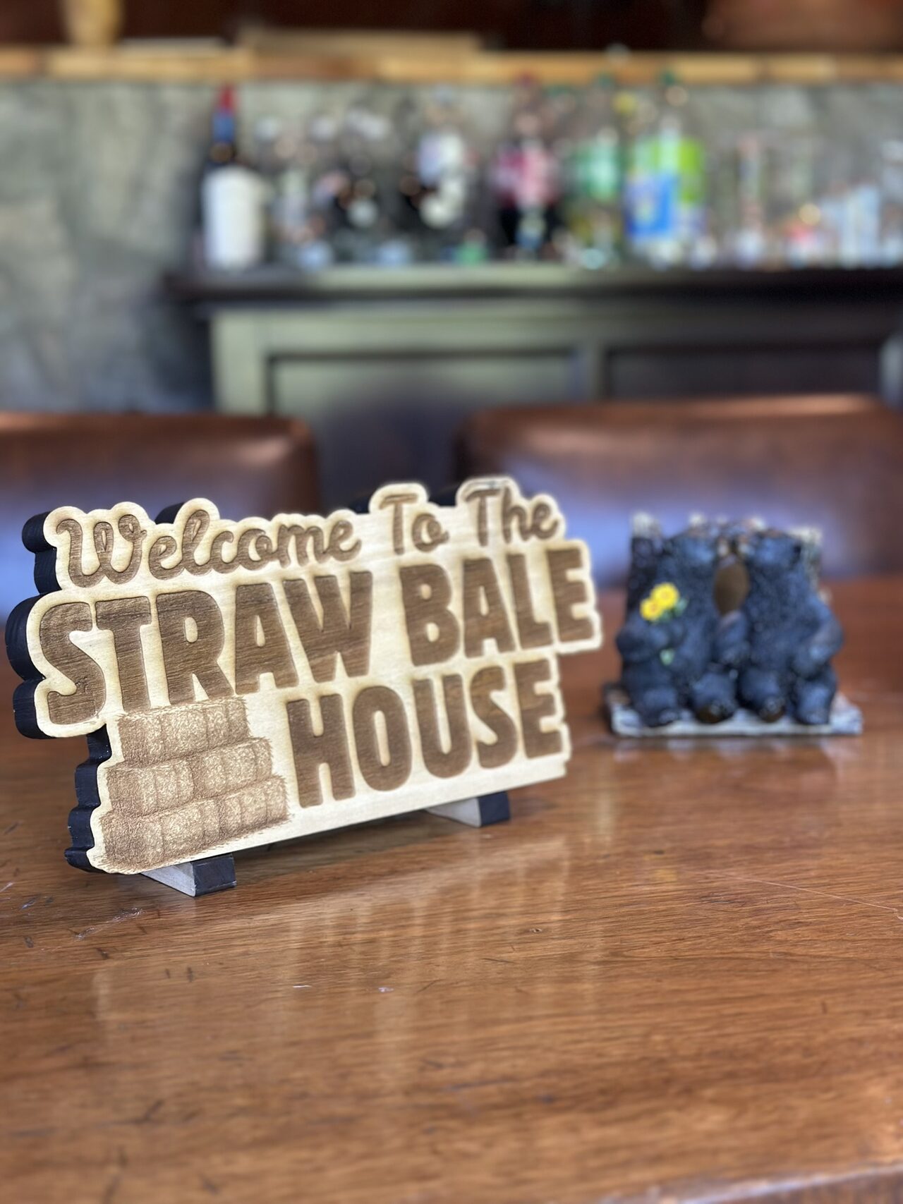 Small wood sign on a polished wooden table reading Welcome To The Straw Bale House in dark engraved lettering, with a stack of engraved straw bales, standing on a small stand with a black bear figurine behind it.