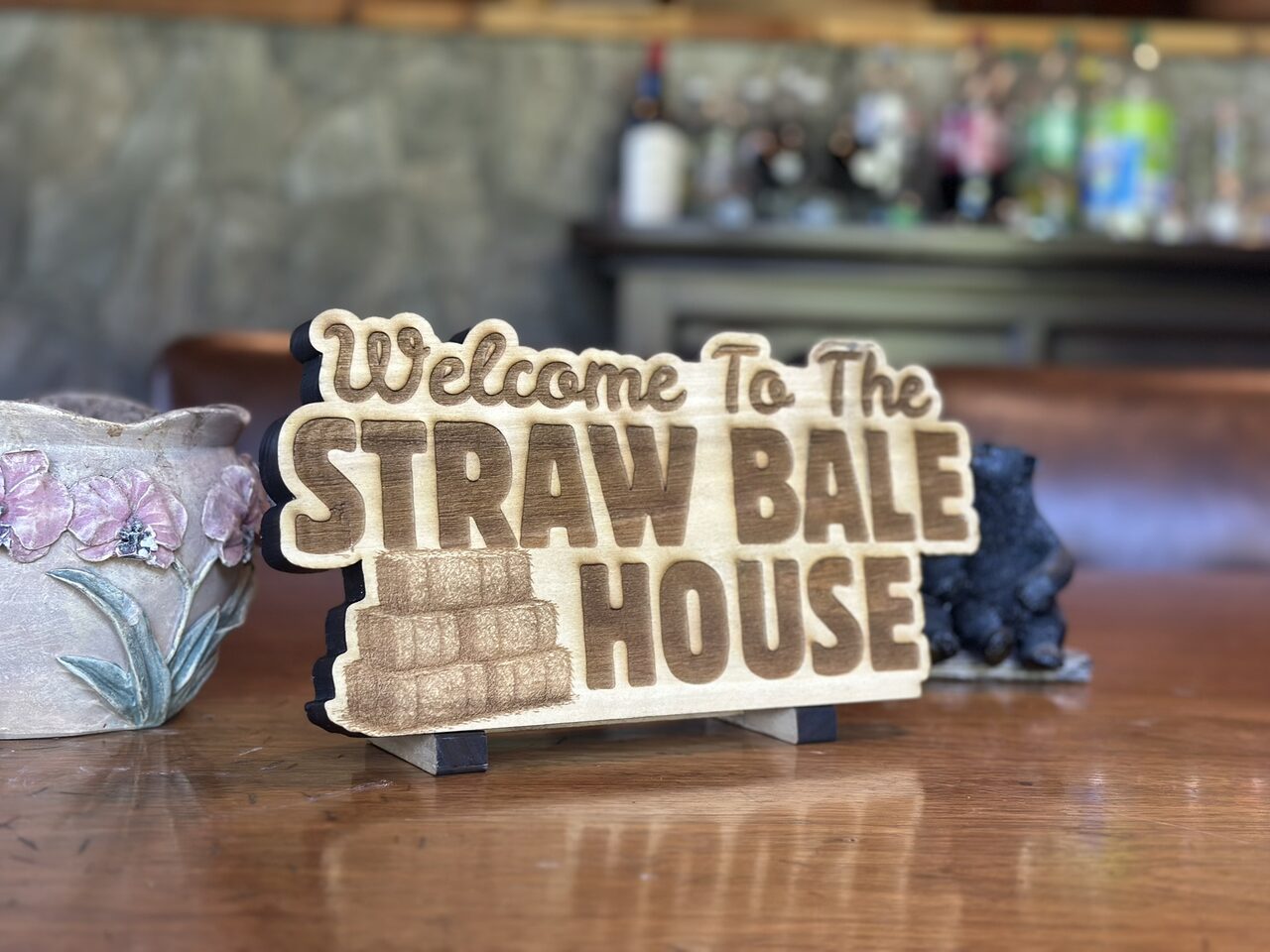 Front view of the engraved wood sign on its stand on a wooden table, with a ceramic bowl painted with pink flowers at the left and a blurred room behind.